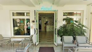 A hotel lobby with a neon sign reading "Hotel," a reception desk, several chairs, and a large potted plant.