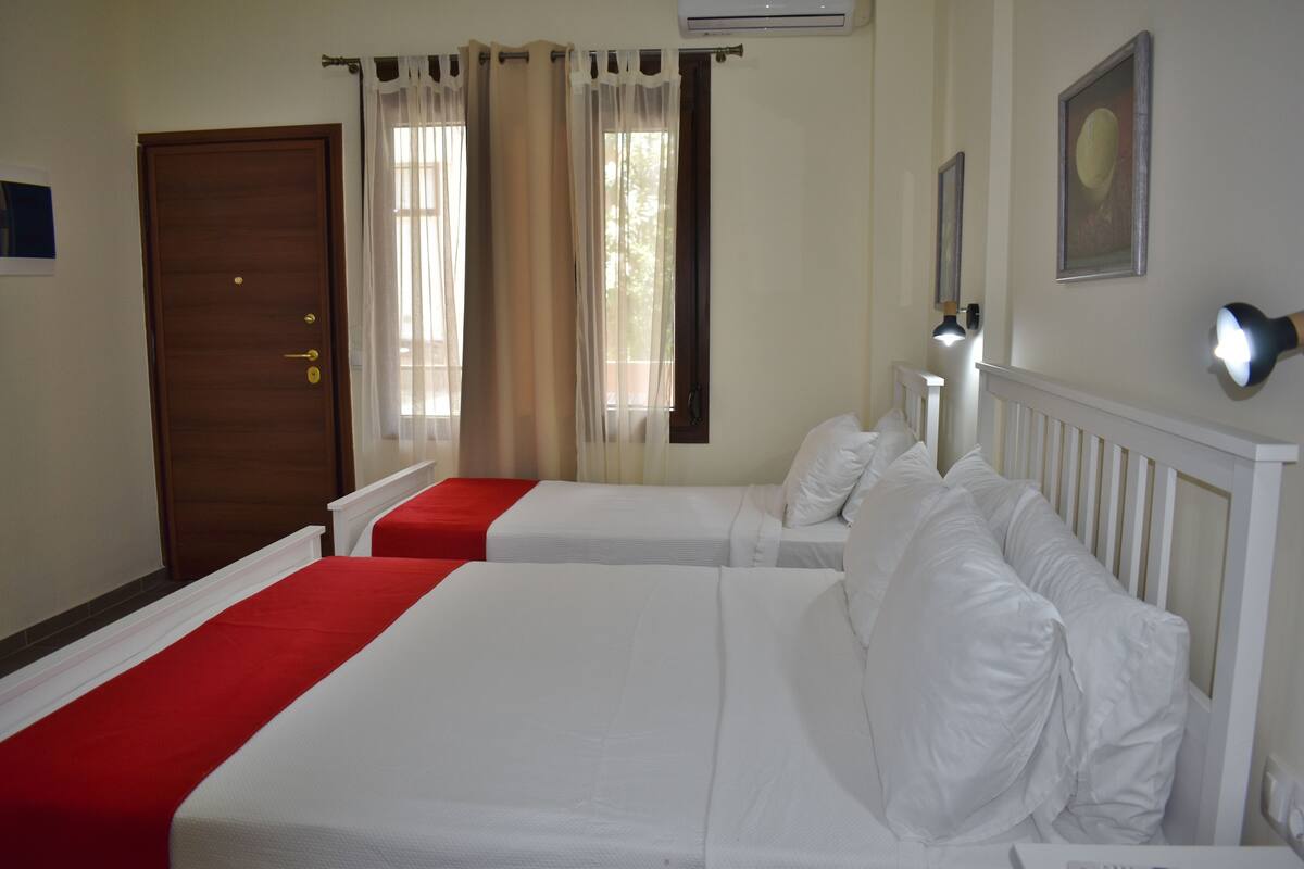 A hotel room with two beds, a wooden door, a window with curtains, and a wall-mounted air conditioner.