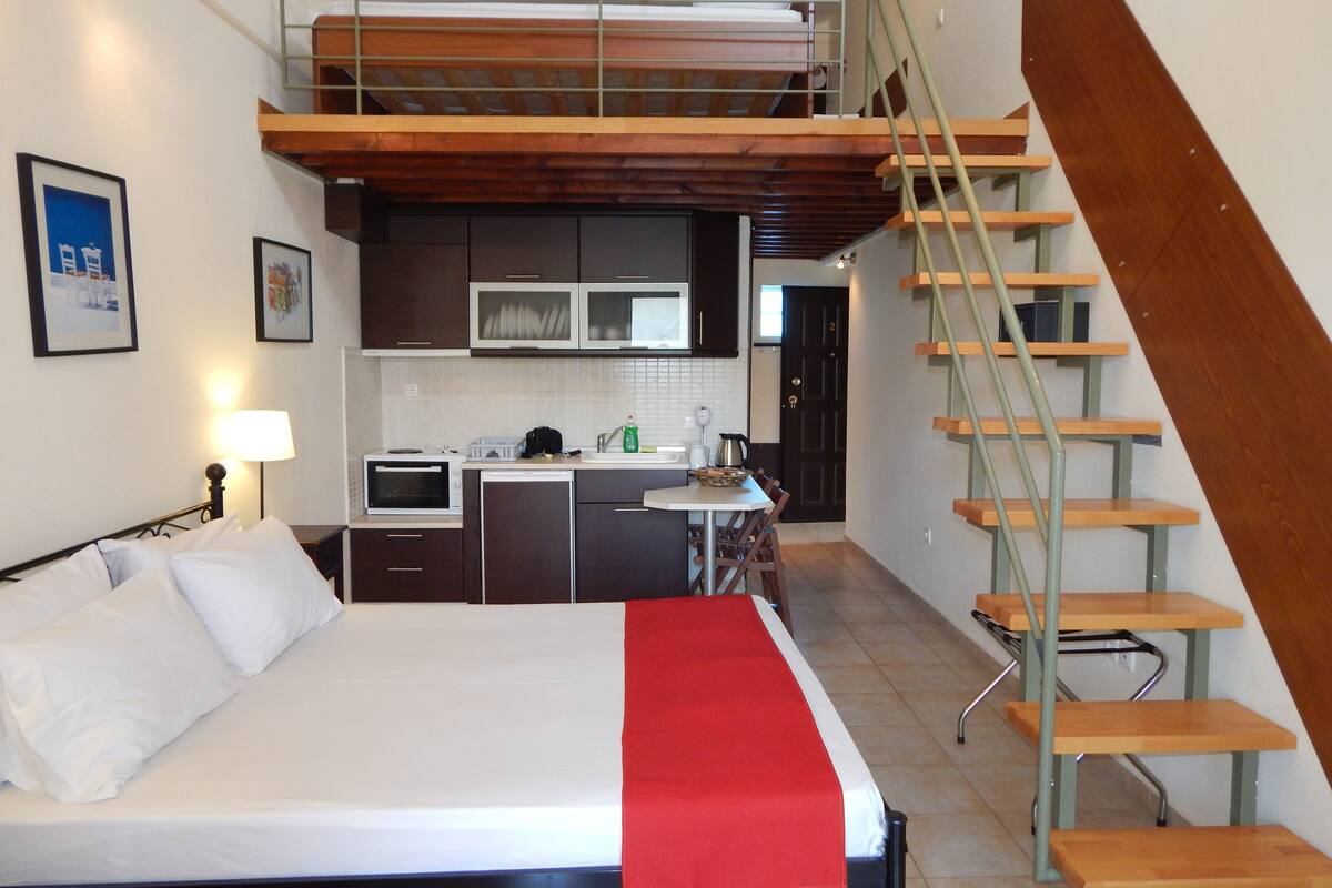 A bedroom with a bed, a red throw, a kitchen with appliances, and a staircase.