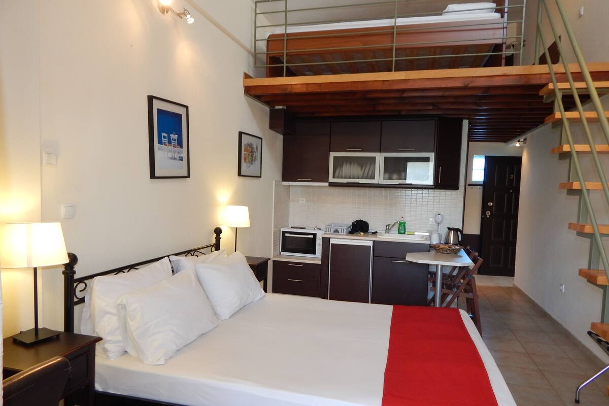 A bedroom with a bed, bedside tables, a desk, a chair, a kitchenette with a microwave, a sink, and cabinets. There is a staircase leading to an upper level.