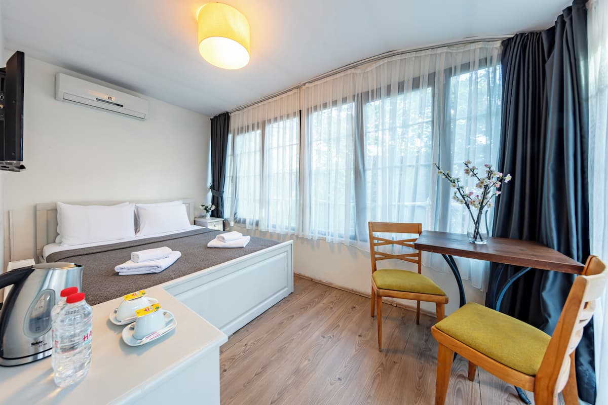 A hotel room with a bed, a table with chairs, a kettle, a bottle of water, and a vase with flowers.