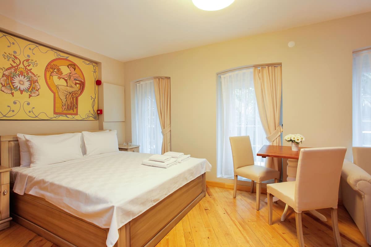 A hotel room with a large bed, a wooden nightstand, a desk with a chair, and a window with curtains.