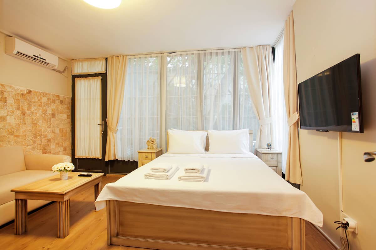 A hotel room with a bed, a small table with a vase of flowers, a nightstand, a TV mounted on the wall, and a window with curtains.