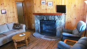 A cozy living room with a stone fireplace, wooden walls, a sofa, and a chair. - Lazy Oaks Resort (Lakeside)