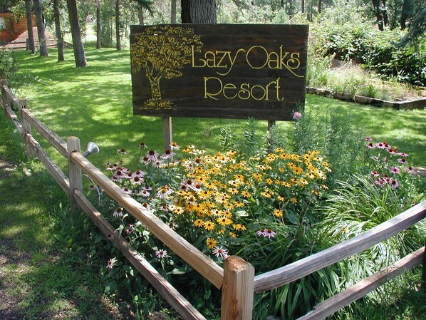 A wooden sign with "Lazy Oaks Resort" written on it, surrounded by a wooden fence and a garden with various flowers.