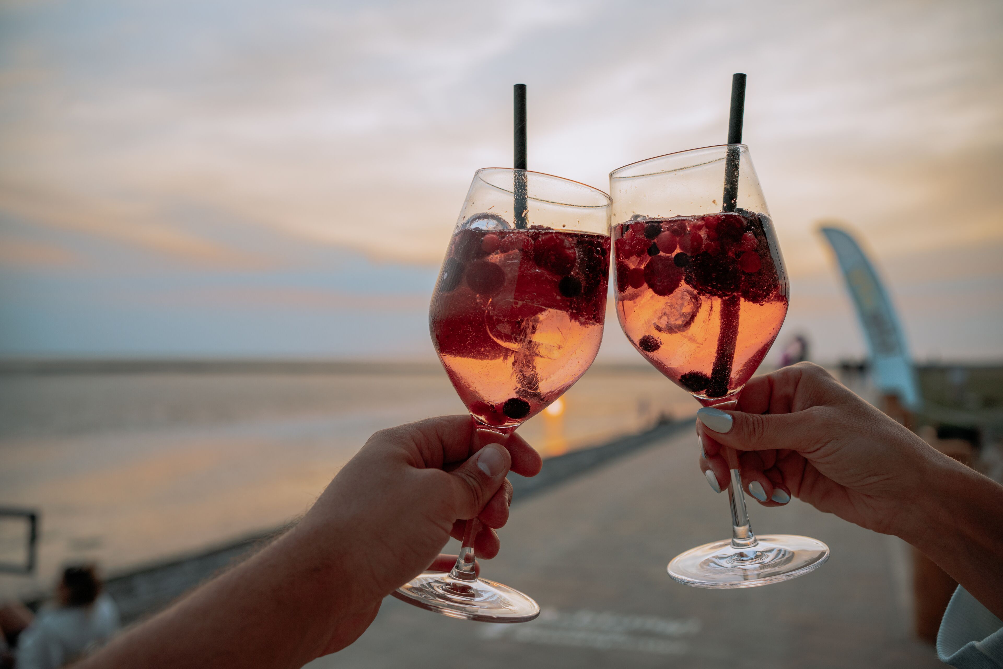 Dos personas sosteniendo vasos de una bebida roja con cubitos de hielo y bayas, con un atardecer y agua de fondo.