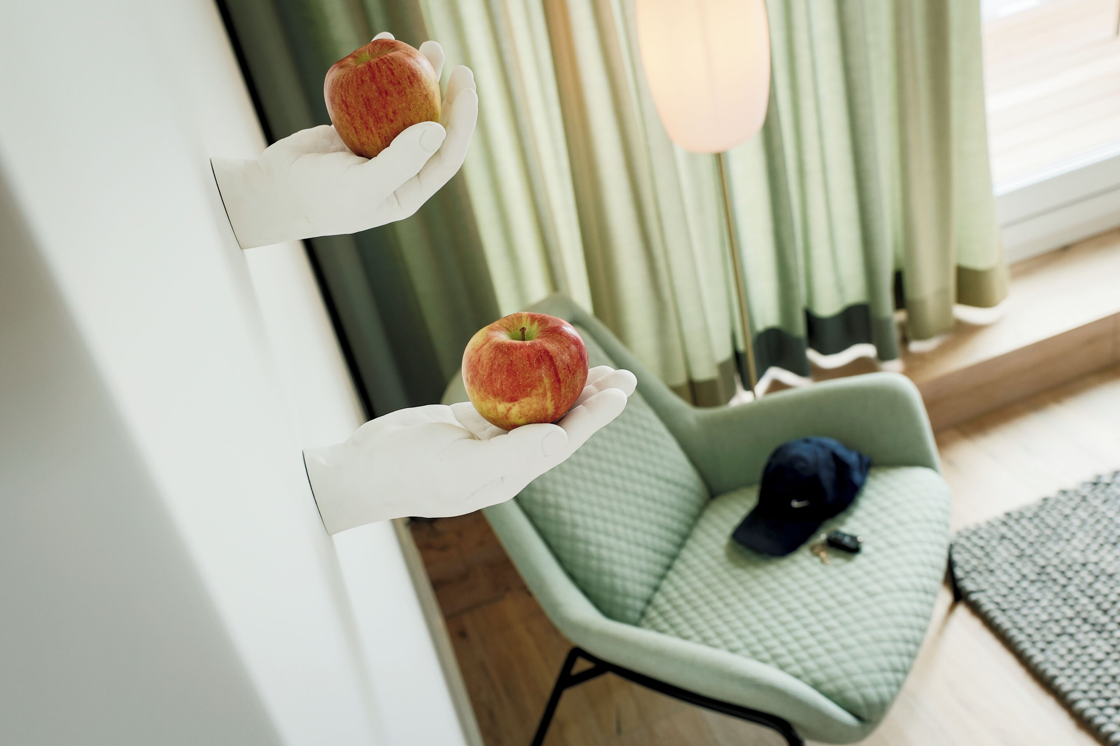 A room with a green armchair, a white wall-mounted shelf with two apples, and a window with curtains.