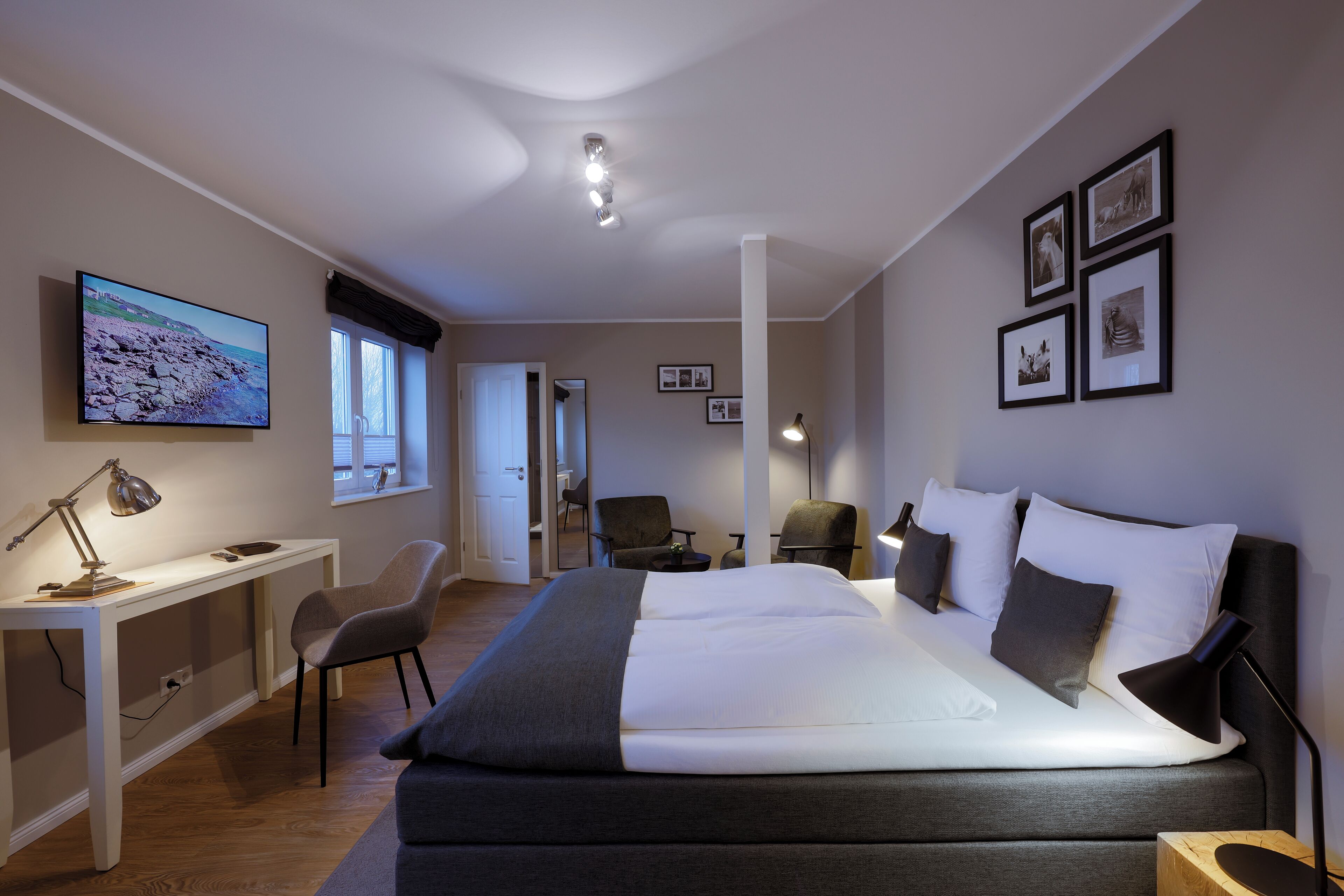 Ein modernes Hotelzimmer mit einem großen Bett, einem Schreibtisch und einem Fernseher. An der Wand hängen gerahmte Bilder und in der Ecke steht ein Sessel.