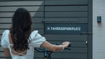 Eine Frau in einem weißen Kleid fährt mit einem Fahrrad vor einem Gebäude, an dem ein Schild mit der Aufschrift 'FAHRRADPARKPLATZ' hängt.