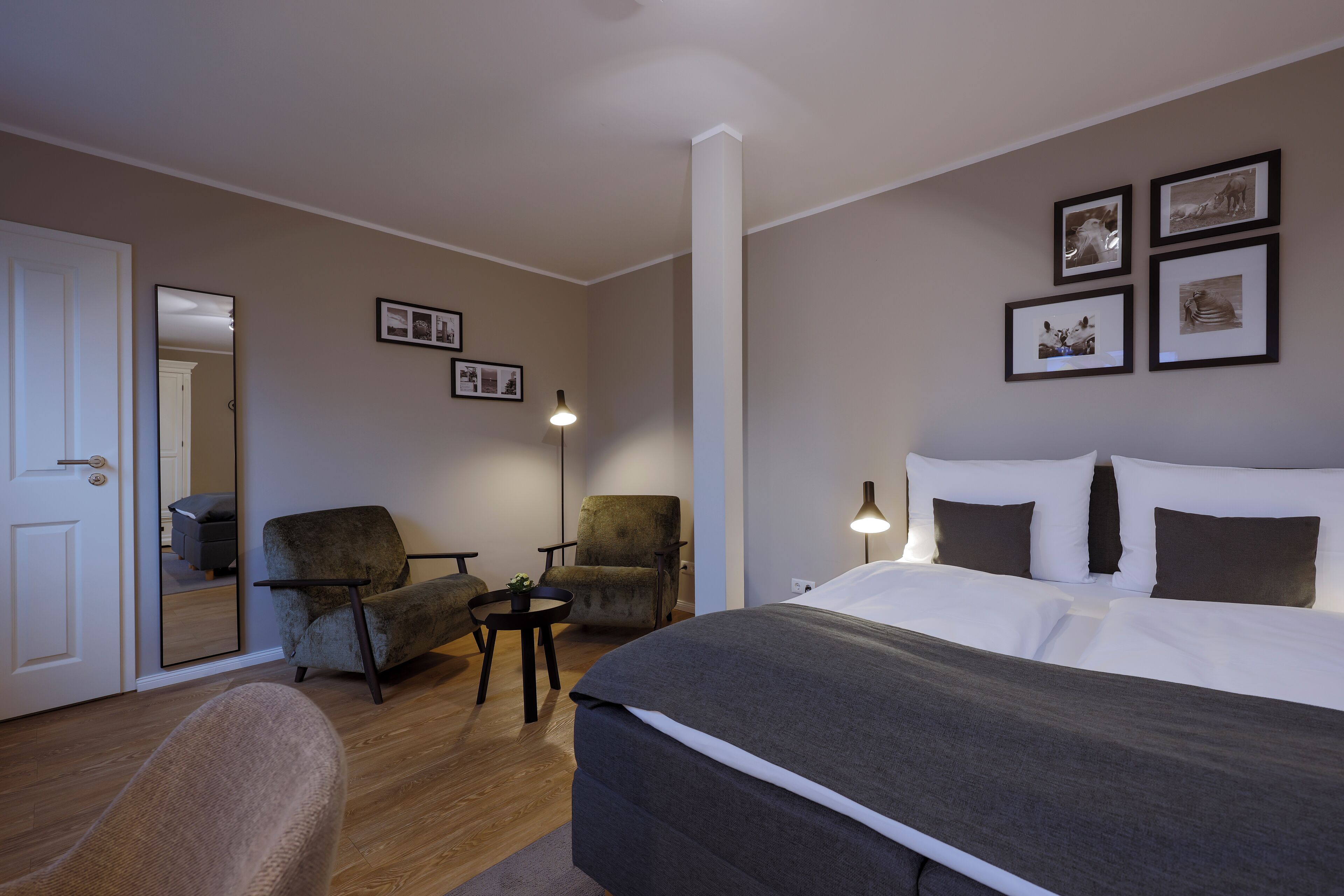 A hotel room with a bed, two armchairs, a small table, and a lamp. There are framed pictures on the wall.