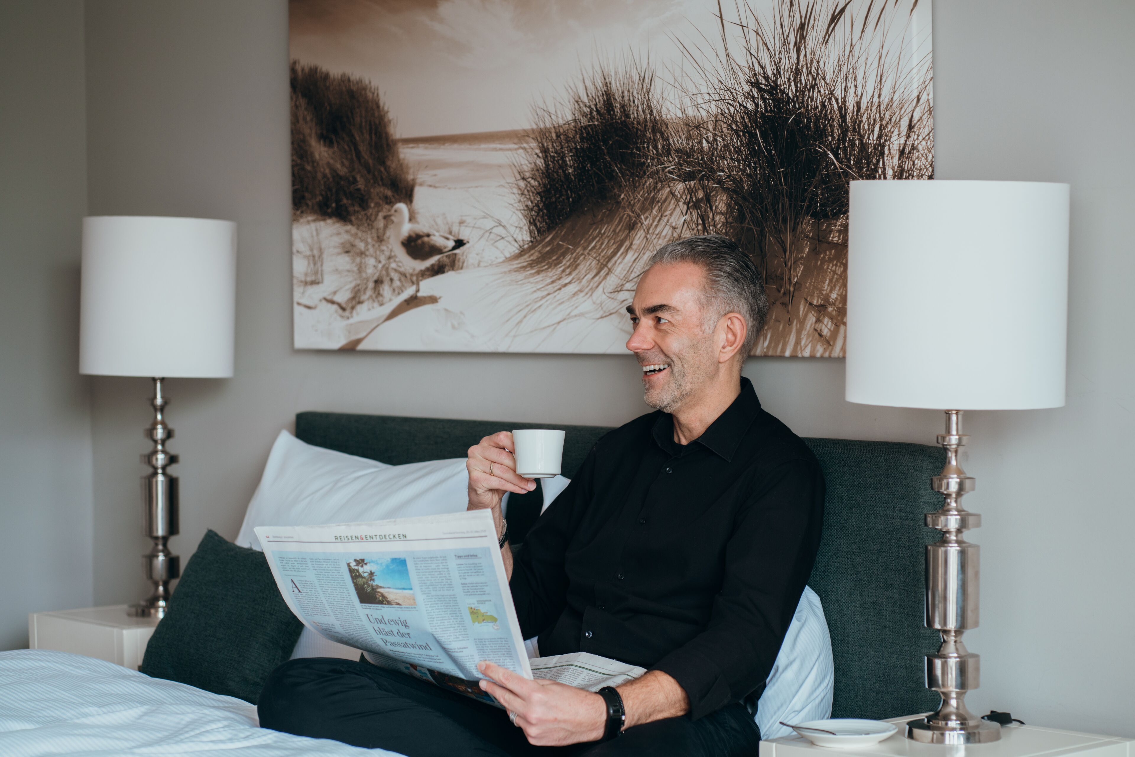 Um homem lê um jornal em um quarto com cama, duas luminárias e um quadro na parede.