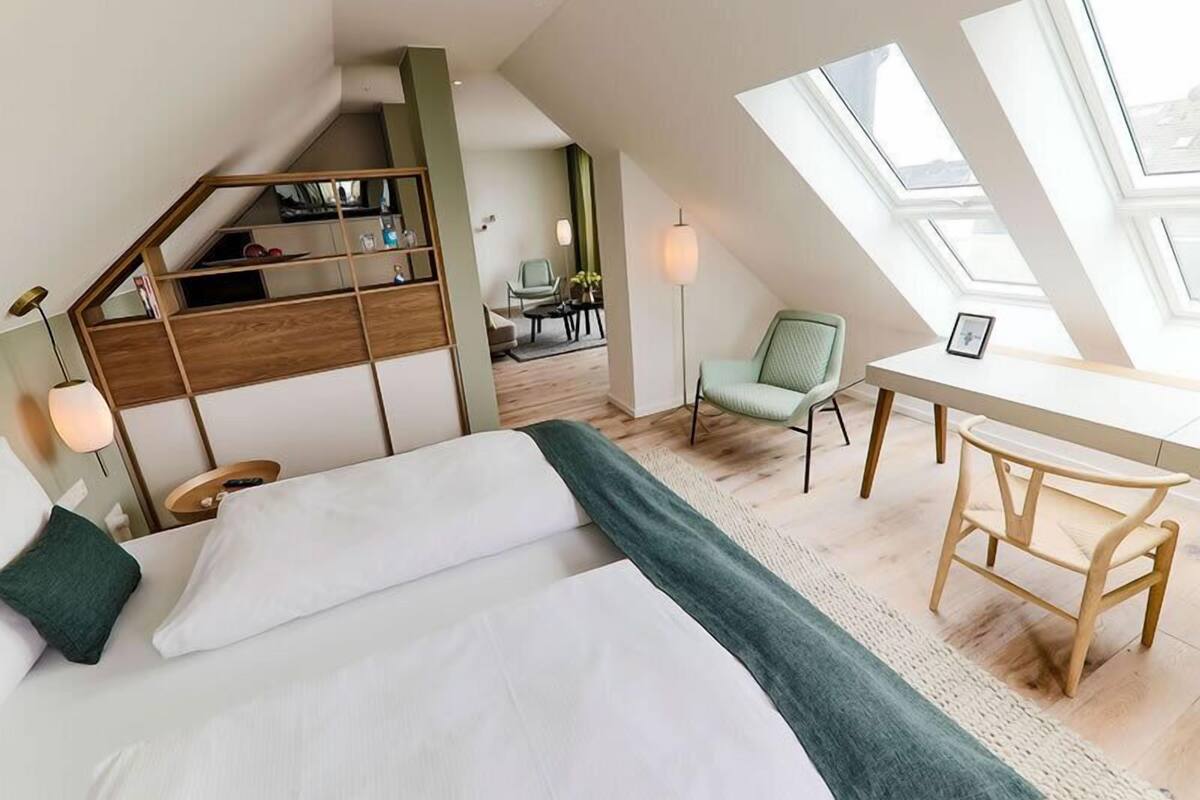 A modern loft bedroom with a large bed, a desk, and a chair. There is a staircase leading to an upper level with a bookshelf.