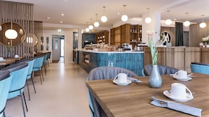 A modern dining area with a wooden table set for two, blue upholstered chairs, and a vase with flowers. - Hotel Astra Maris  (Büsum)