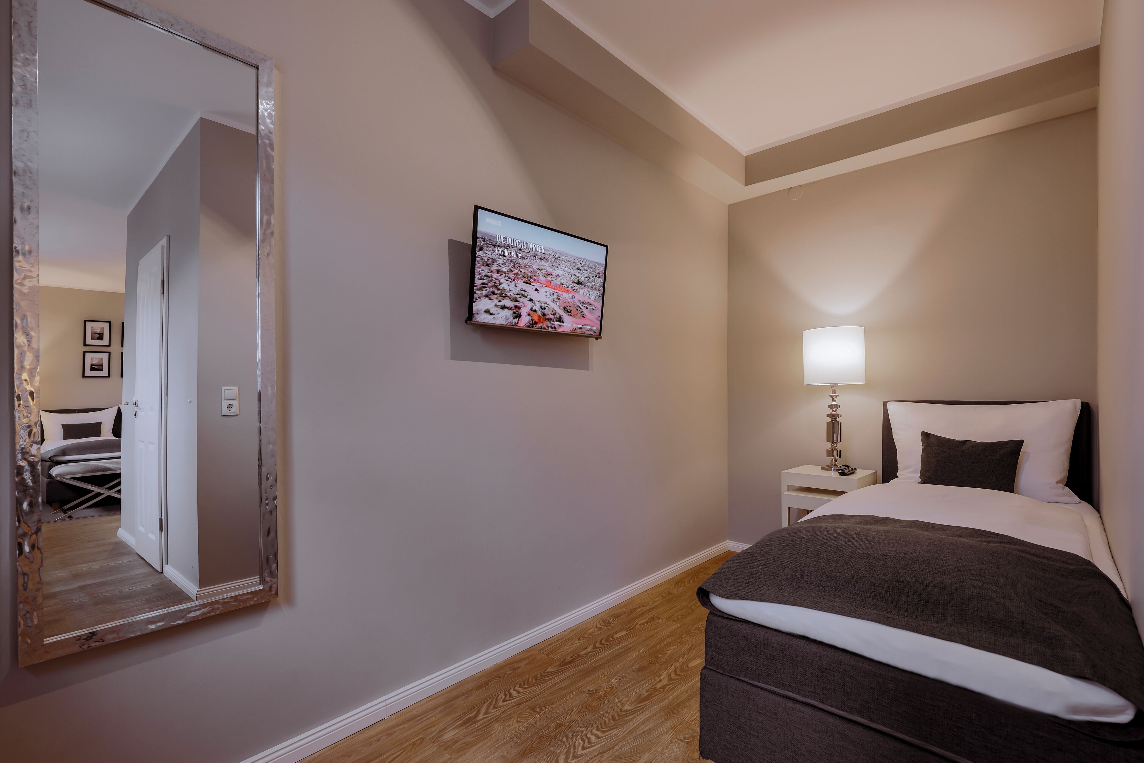 Habitación de hotel con un espejo grande, un televisor montado en la pared, una mesita de noche con lámpara y una cama bien hecha.