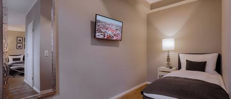 A hotel room with a large mirror, a television mounted on the wall, a bedside table with a lamp, and a neatly made bed.