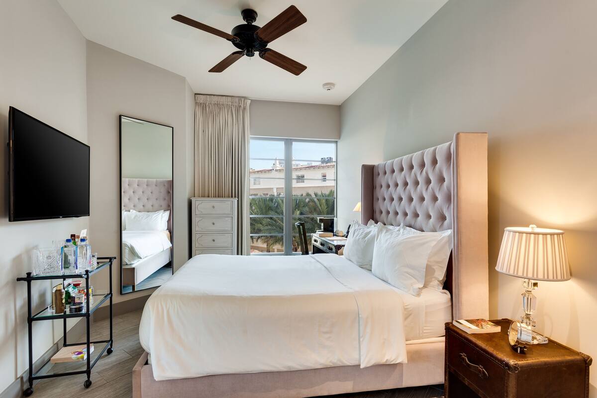 A hotel room with a large bed, a ceiling fan, a TV, a window with a view, and a bedside table with a lamp.