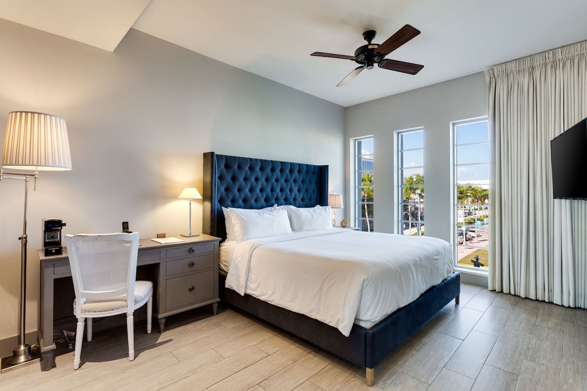 A bedroom with a bed, a desk, a chair, a ceiling fan, and large windows.