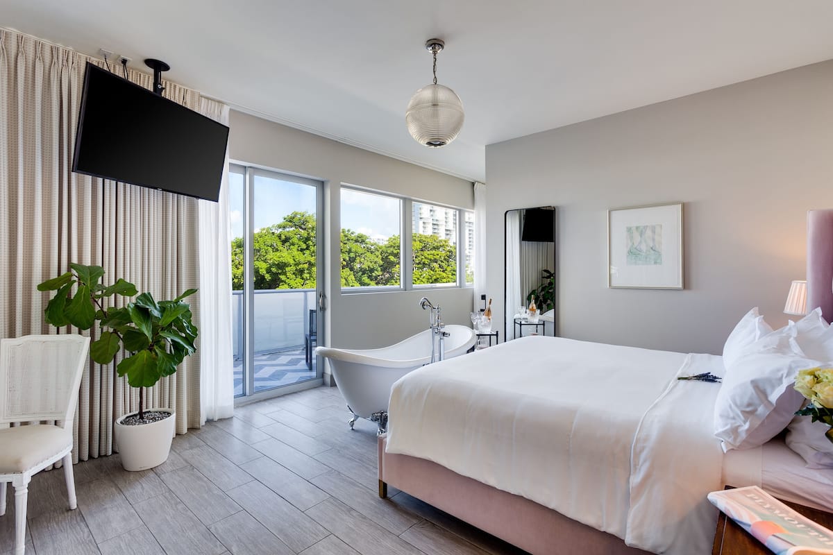 A modern hotel room with a large bed, a bathtub, a flat-screen TV, and a balcony with a view of trees and buildings.