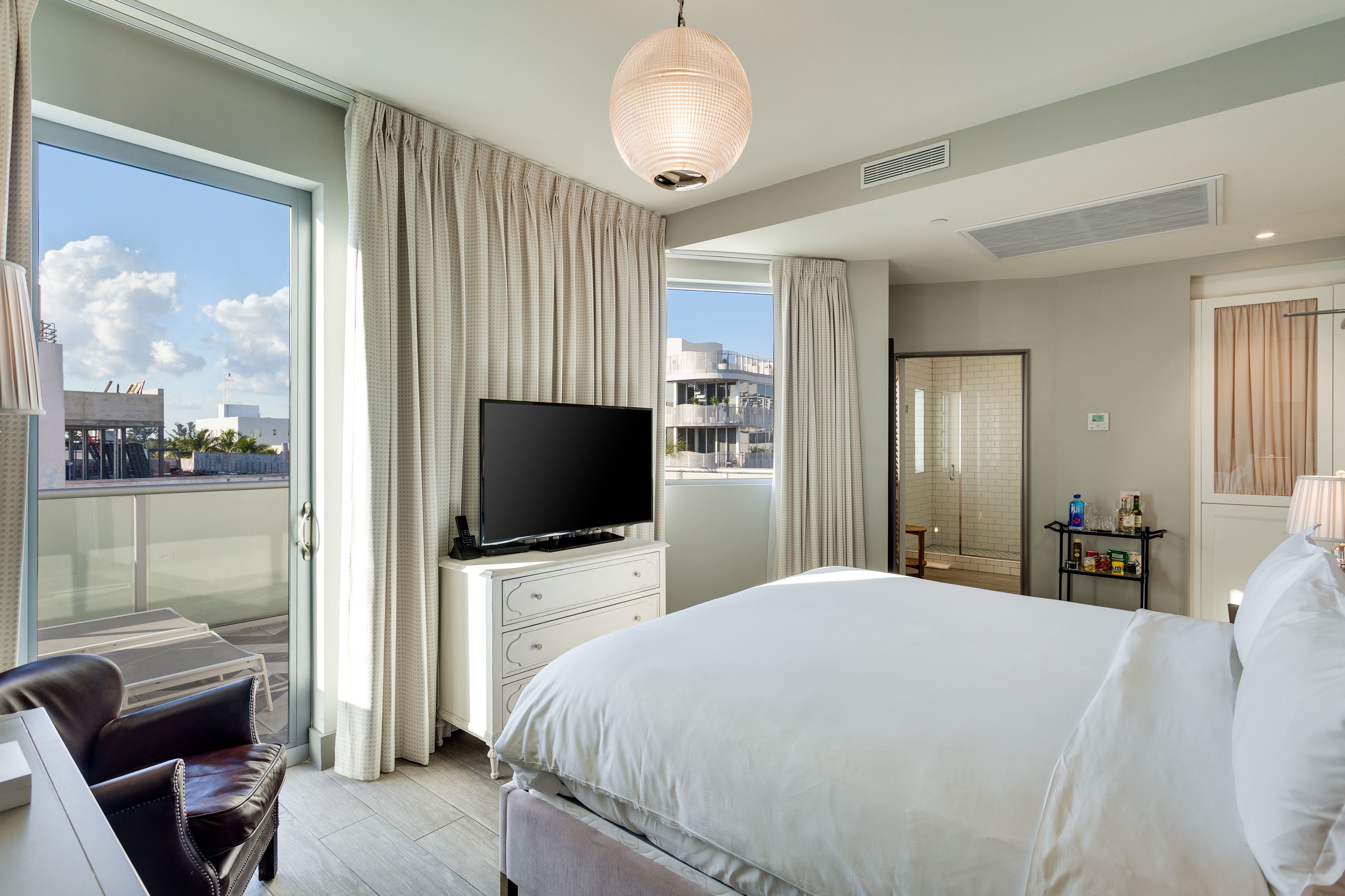 A modern hotel room with a large bed, a flat-screen TV on a white dresser, a balcony with a glass door, and a view of the cityscape.
