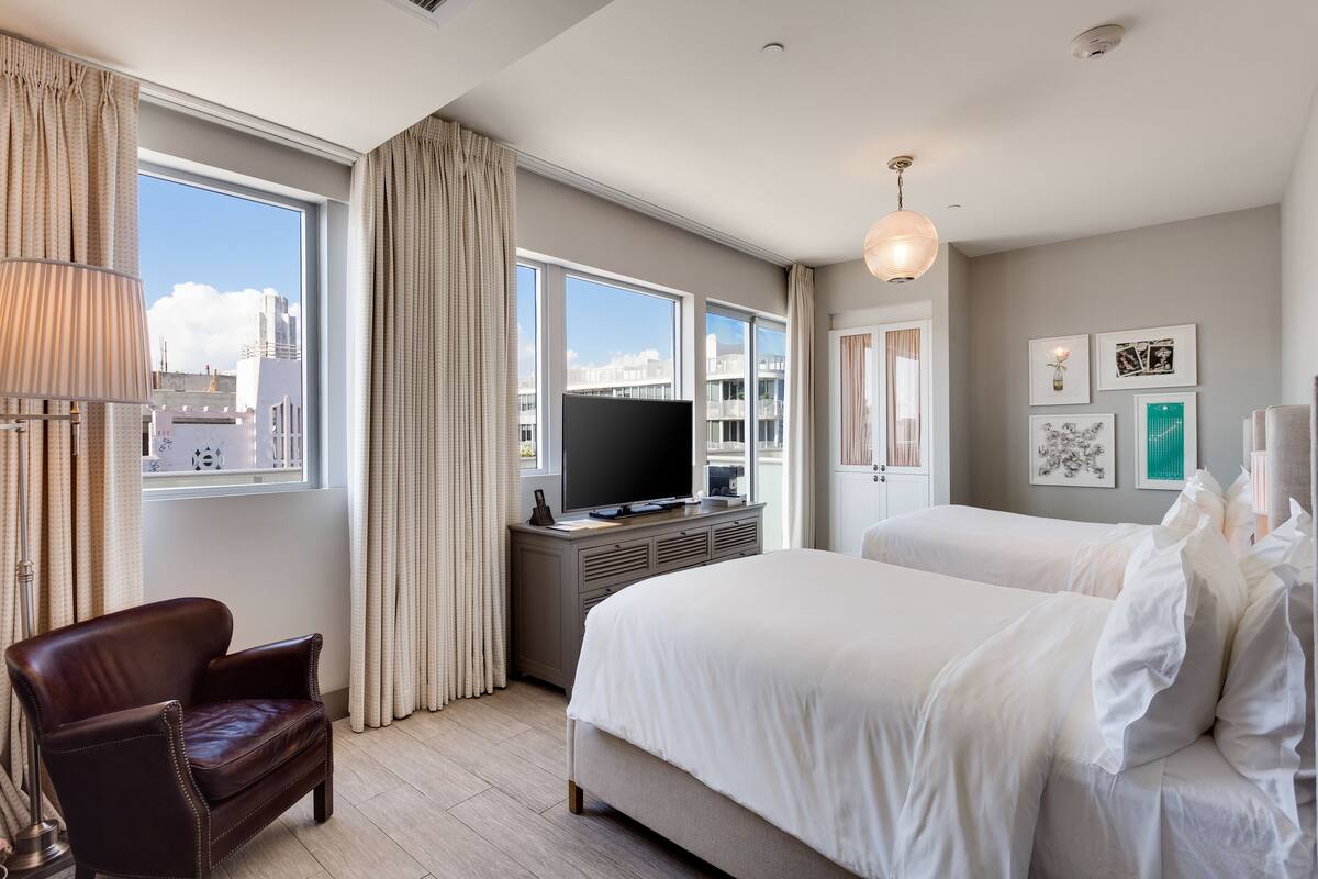 A hotel room with a large bed, a TV on a stand, a chair, and a window with curtains.