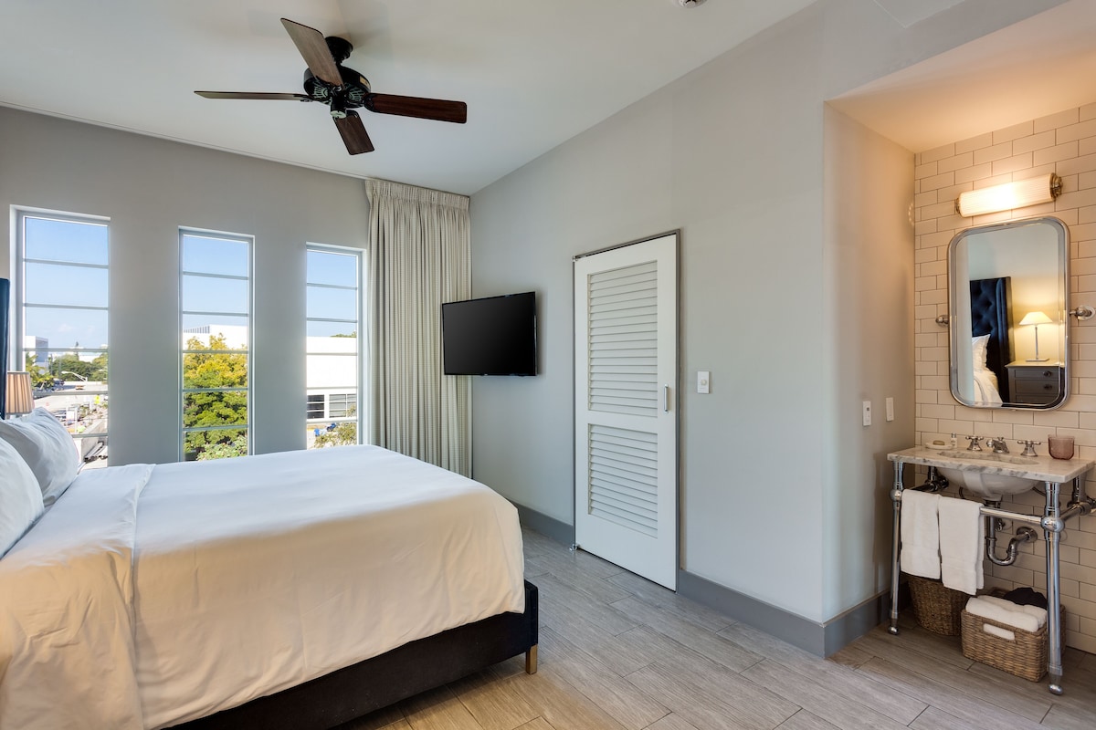 A bedroom with a bed, a ceiling fan, a TV, a window, and a bathroom with a sink and mirror.