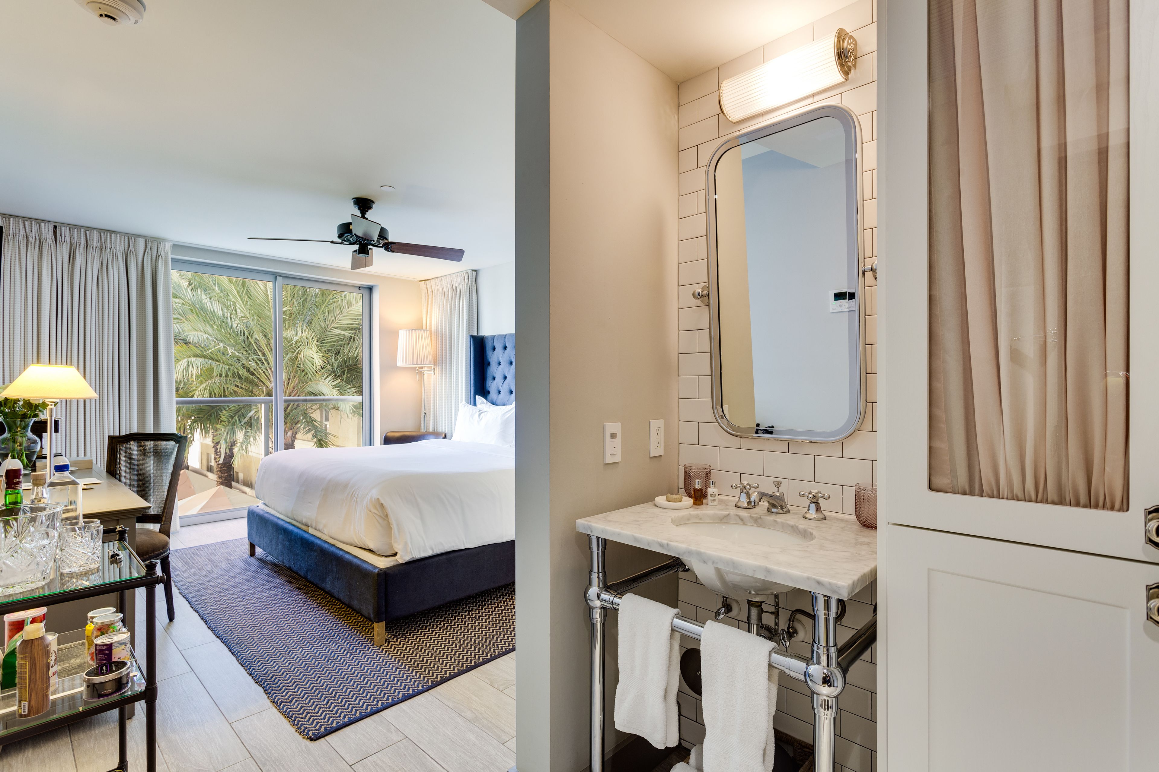 Habitación de hotel con cama, escritorio con lámpara, ventana grande y baño con lavamanos y espejo.