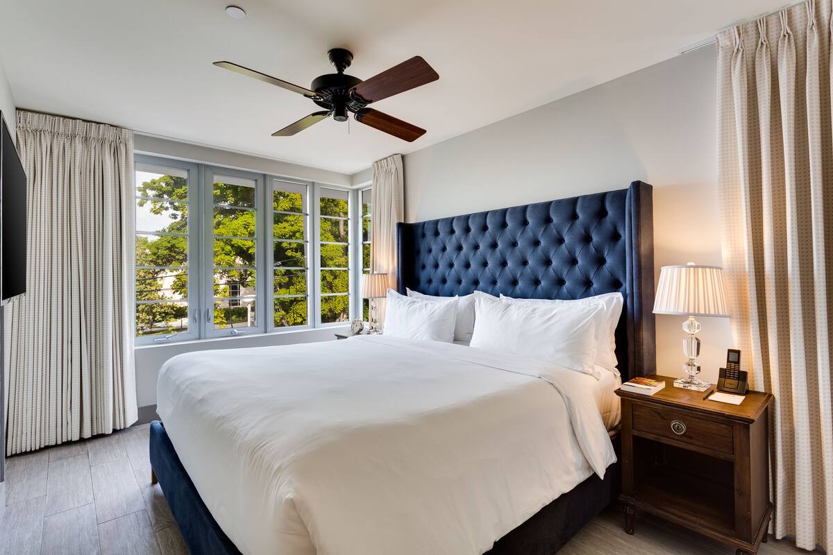 A hotel room with a large bed, a ceiling fan, a nightstand with a lamp, and a view of trees through the window.