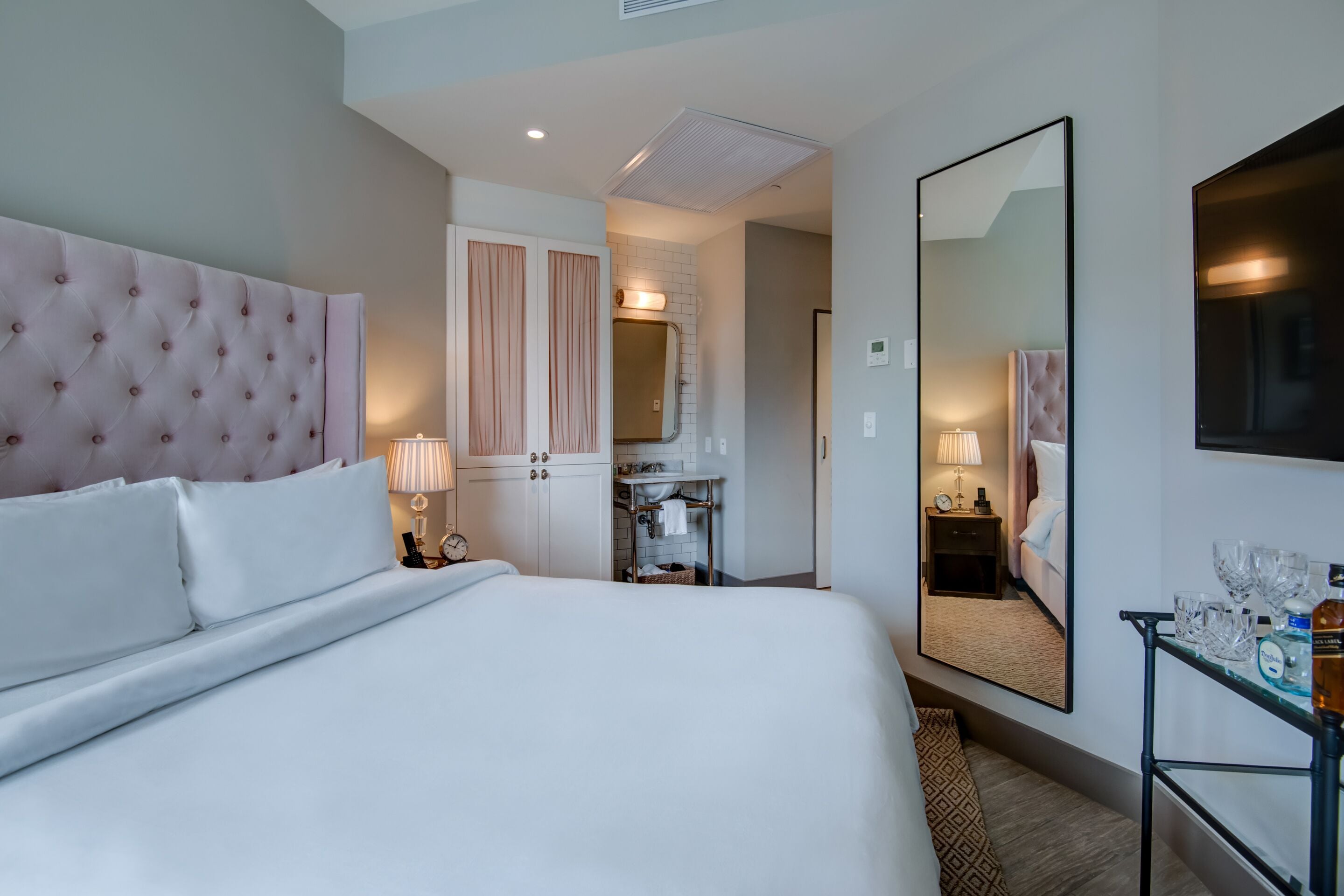 A hotel room with a large bed, a vanity with a mirror, and a small table with glasses and a bottle.
