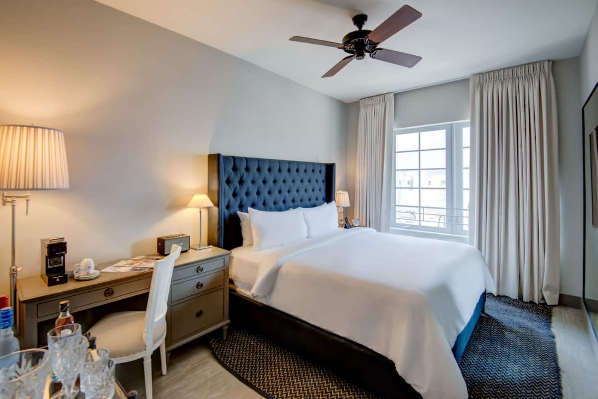 A hotel room with a large bed, a ceiling fan, a desk with a lamp, a chair, and a window with curtains.