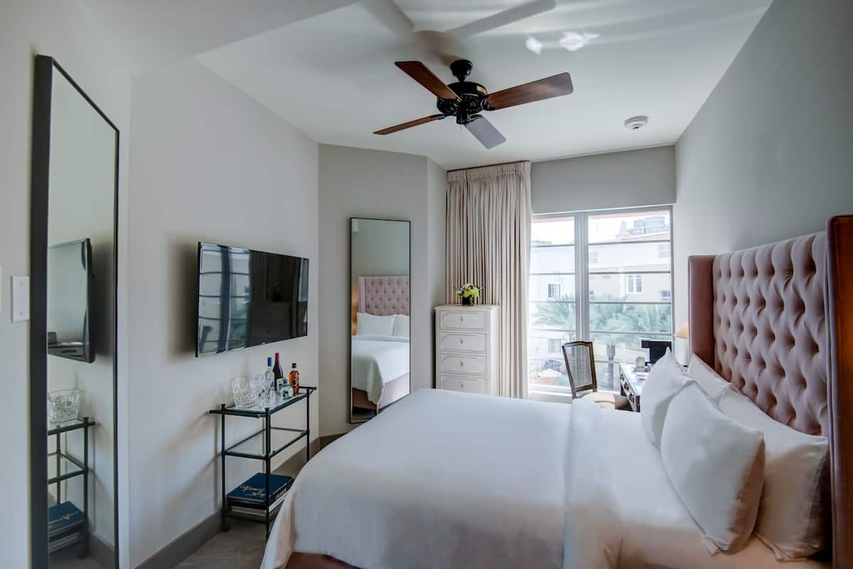 A hotel room with a large bed, a ceiling fan, a TV, a dresser, and a window with a view of buildings.