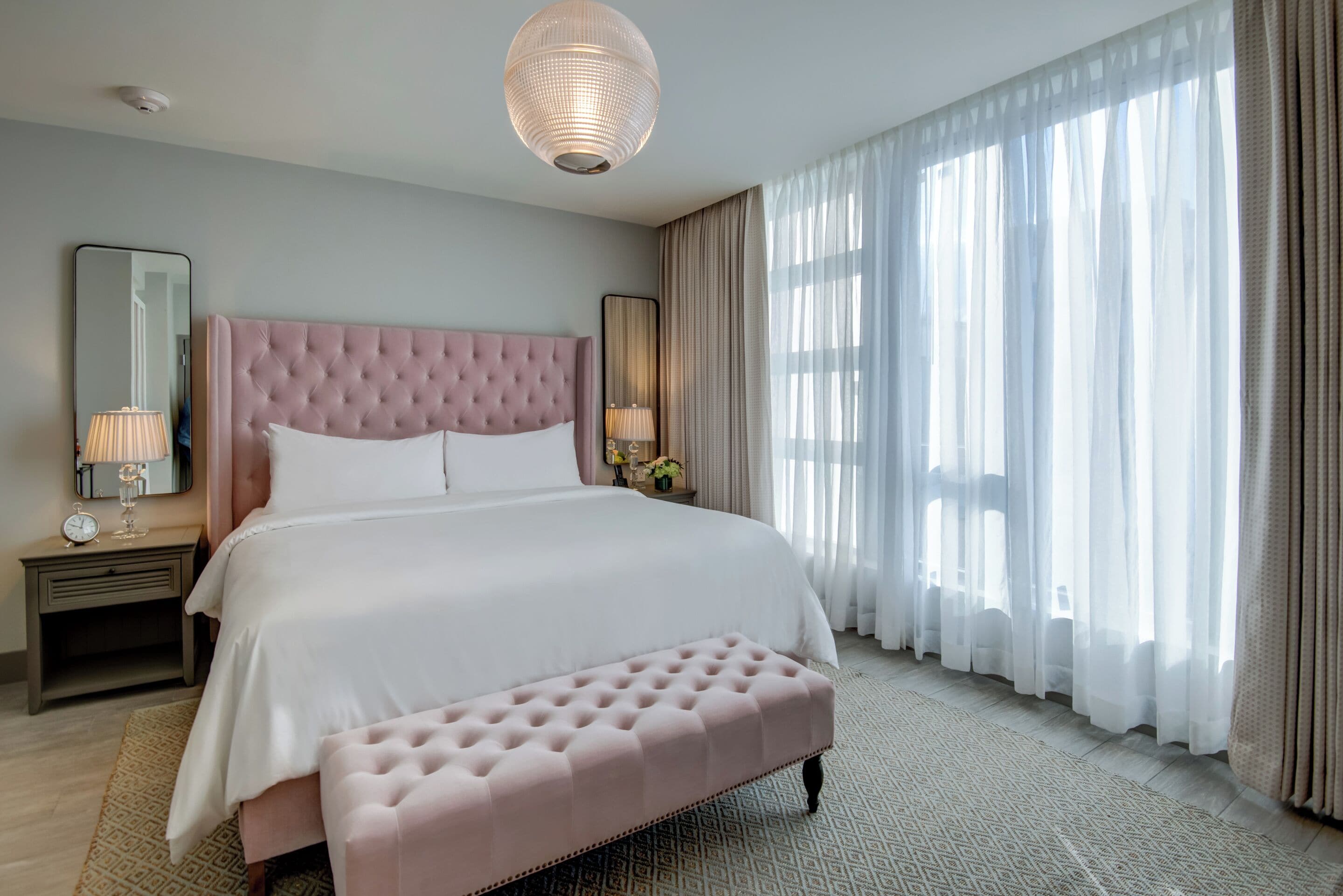 A spacious bedroom with a large bed, a tufted headboard, a bedside table with a lamp, and sheer curtains allowing natural light.