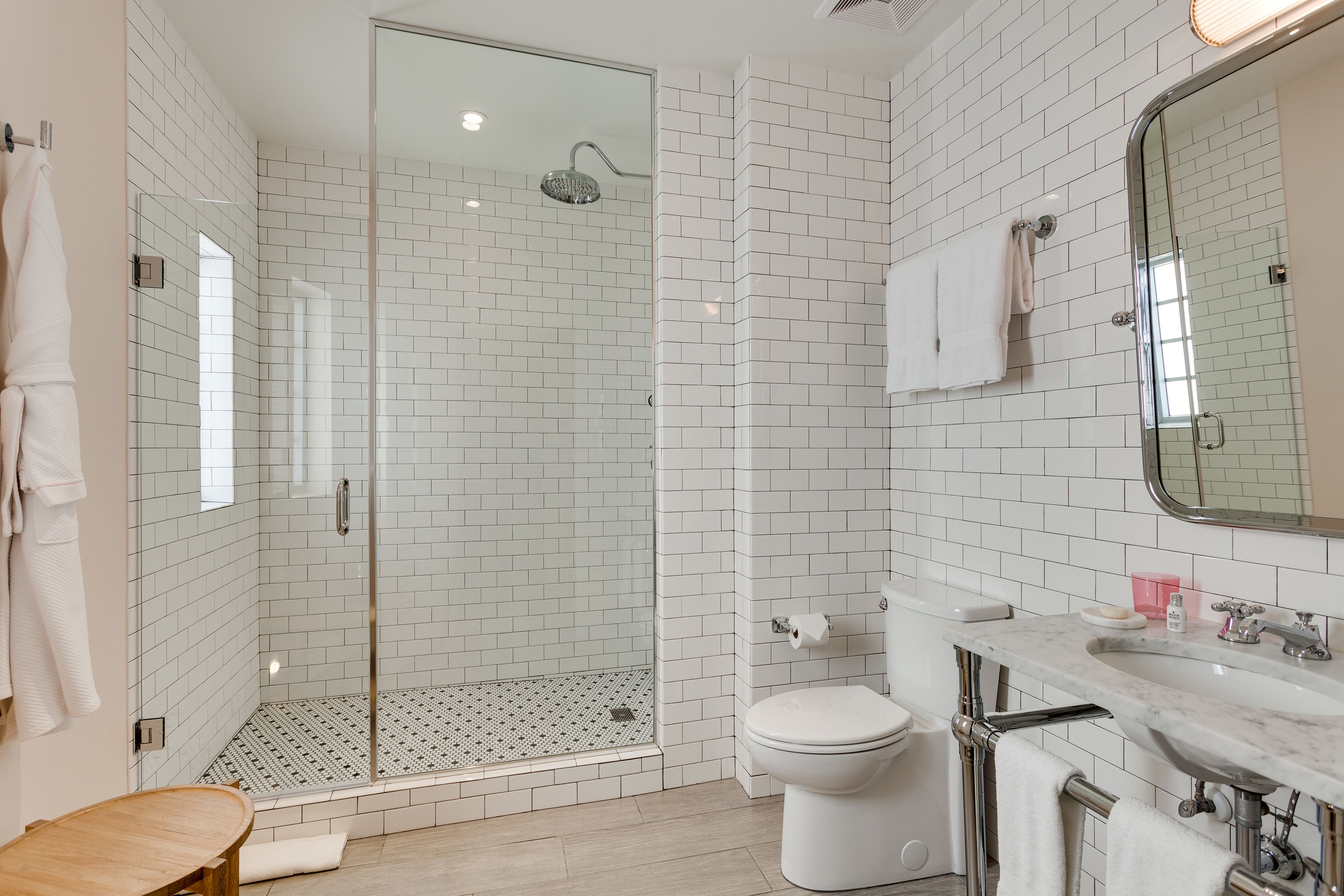 A modern bathroom with a glass-enclosed shower, white tiled walls, a marble sink, and a wooden stool.