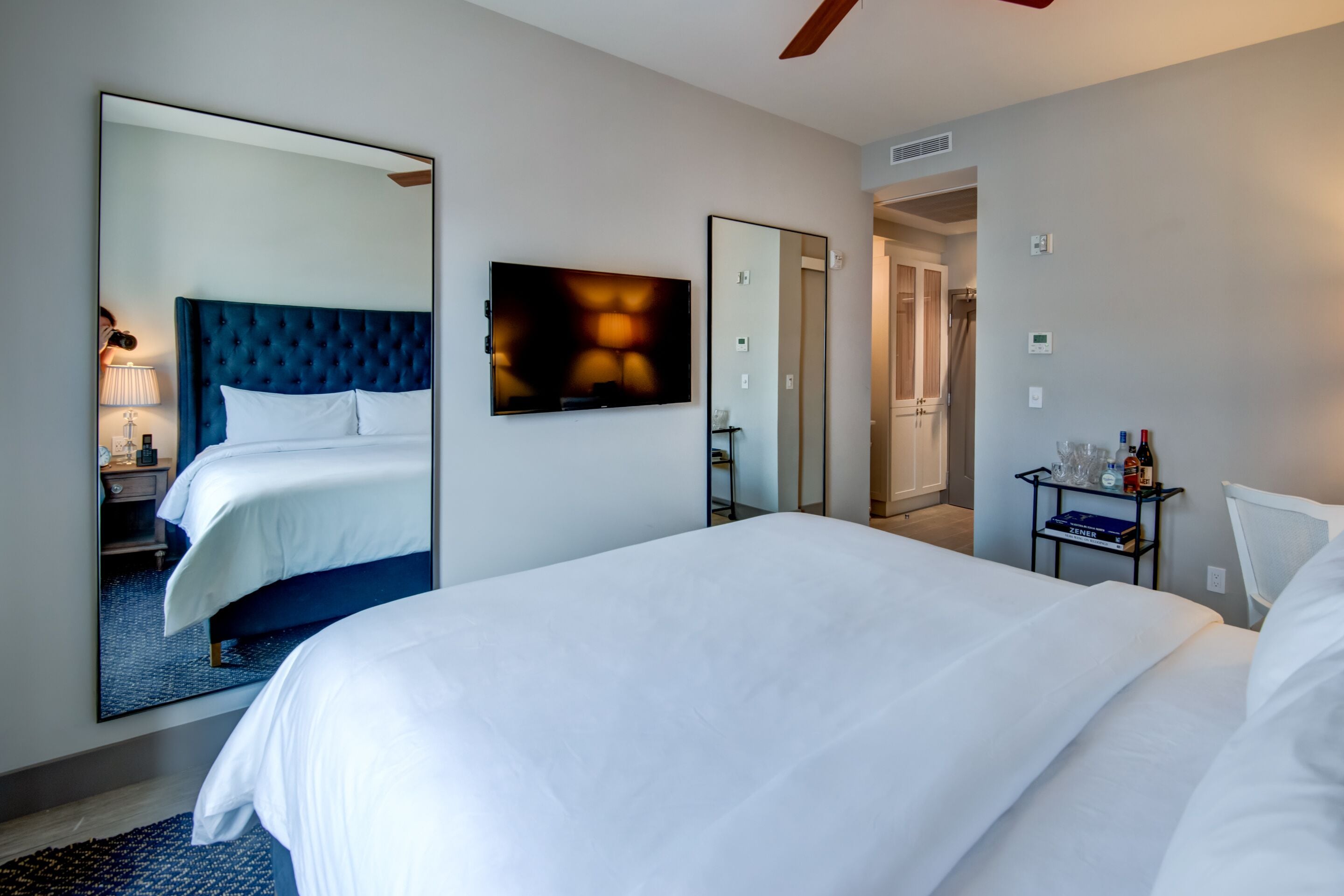 A hotel room with a large bed, a mirror, a bedside table with a lamp, and a small shelf with toiletries.