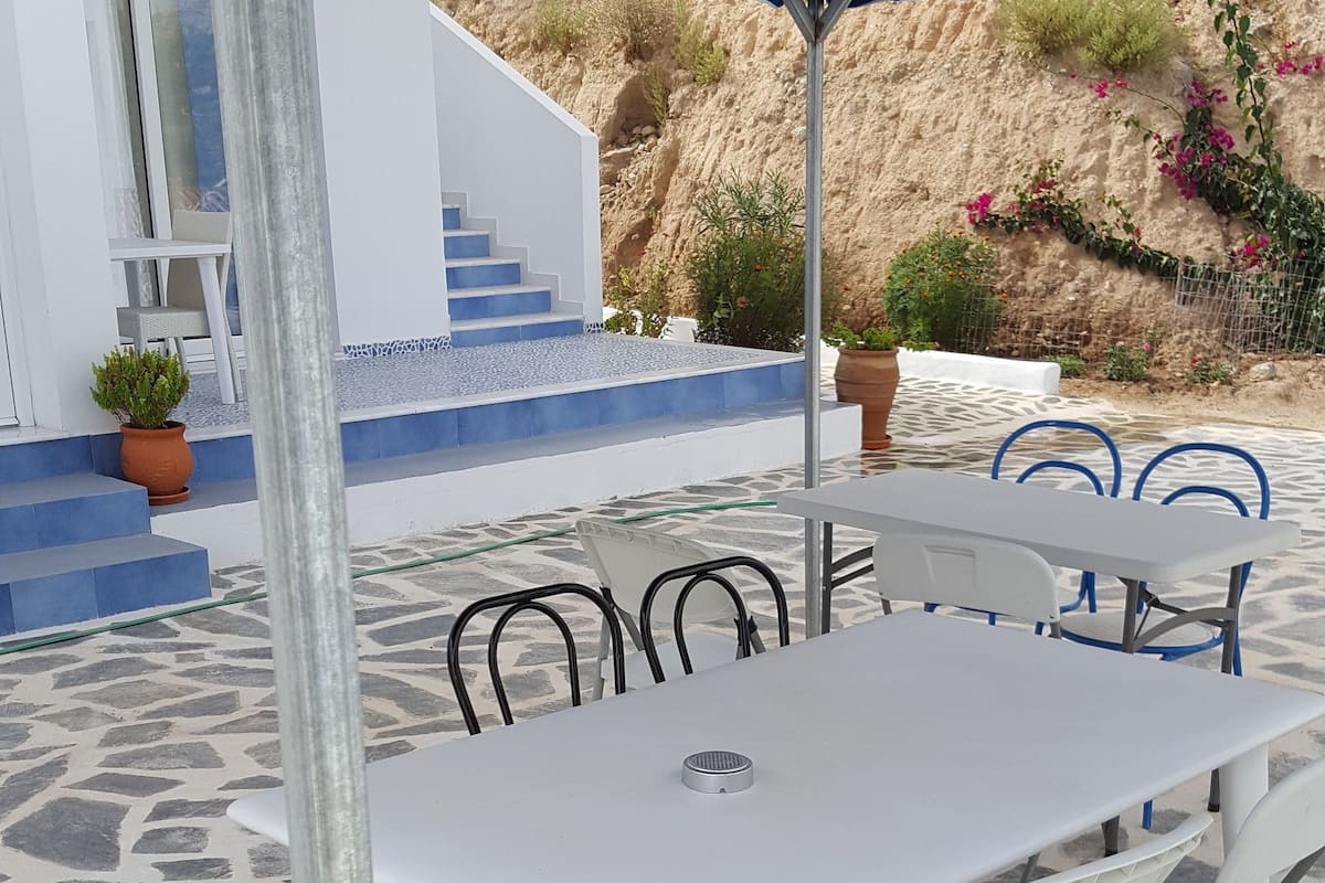 An outdoor seating area with a white table and chairs under a blue canopy, set on a patterned stone floor with a staircase and potted plants in the background.
