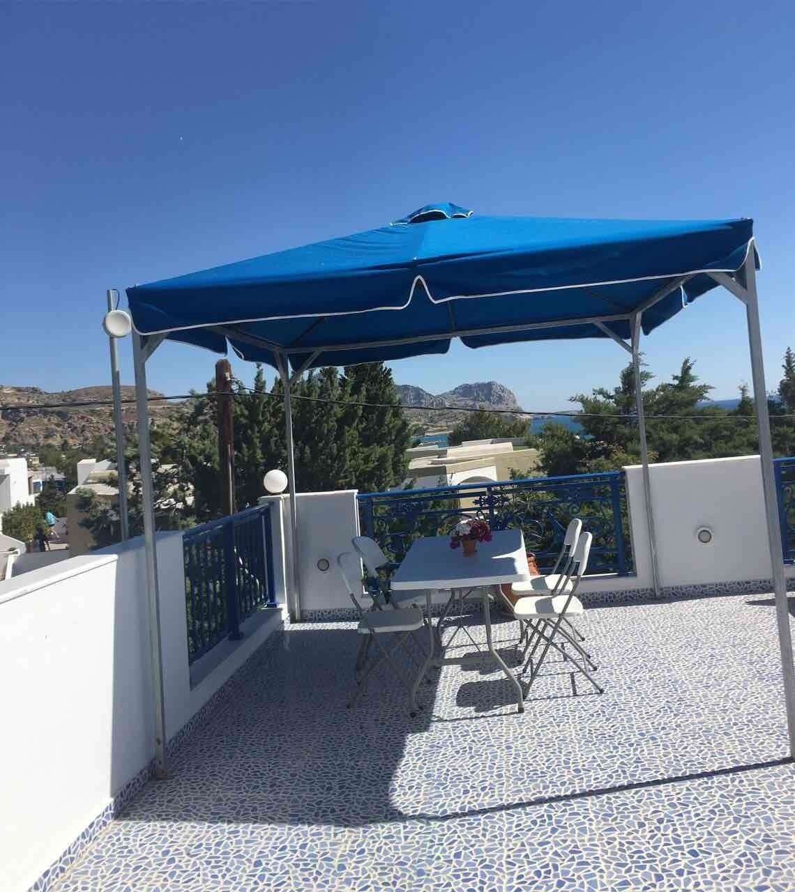 En terrasse med et bord og stole under et blåt markise, med udsigt over et naturskønt landskab.
