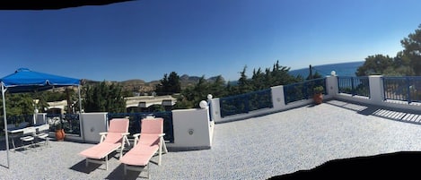 A rooftop terrace with a clear view of the sea, furnished with lounge chairs and a table under a blue canopy.