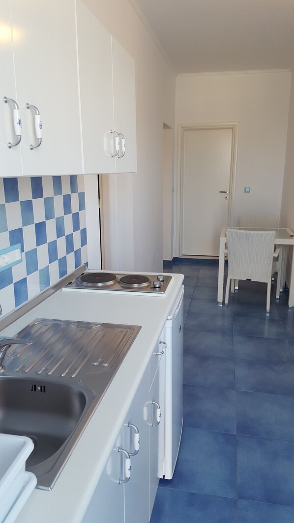 Una cocina con armarios blancos, una estufa y un lavamanos. Se aprecia un salpicadero a cuadros azul y blanco, y una mesa con mantel blanco al fondo.