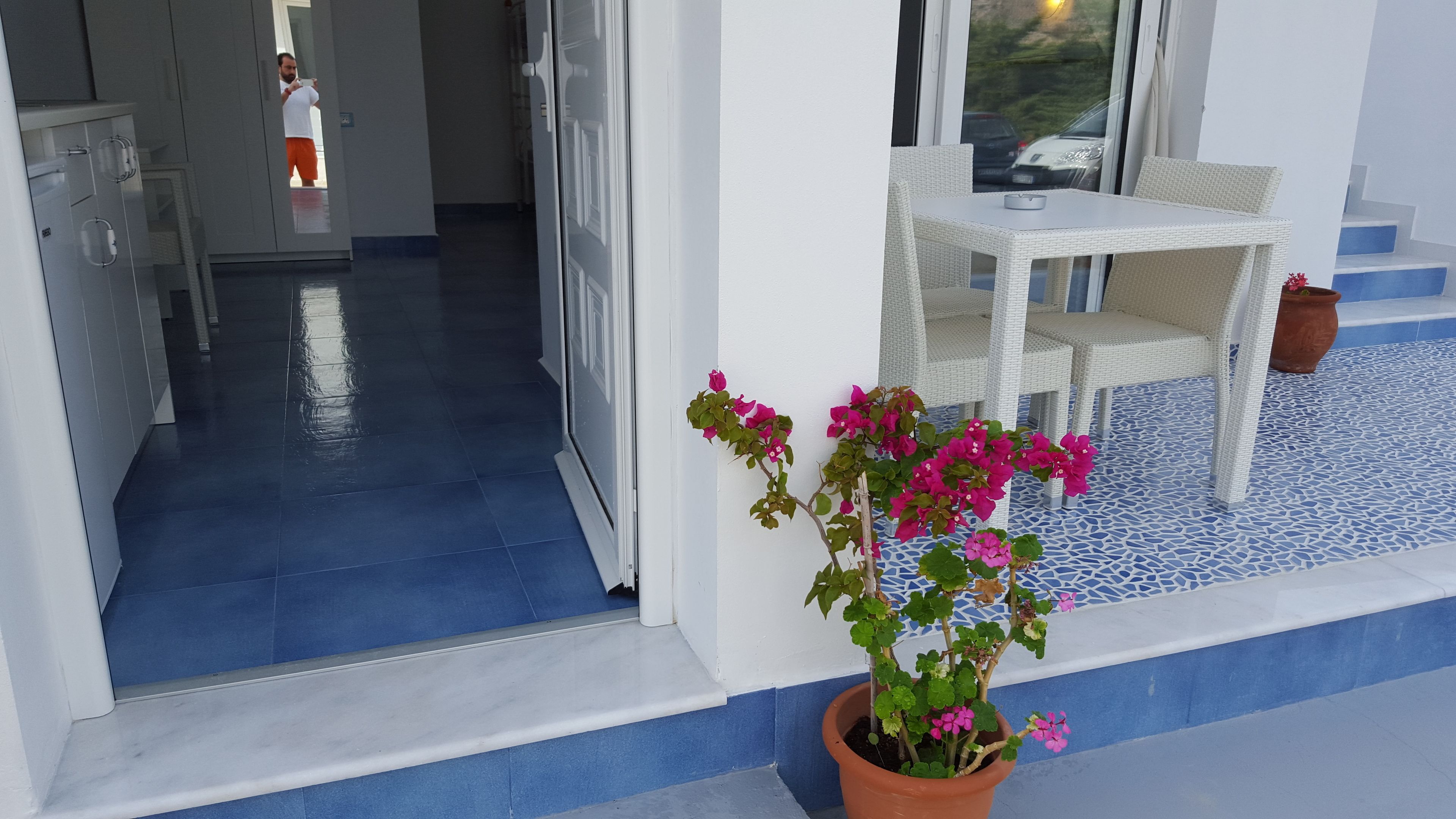 Een hotelentree met een blauw betegelde vloer, een witte tafel en stoelen buiten, en een potplant met roze bloemen.