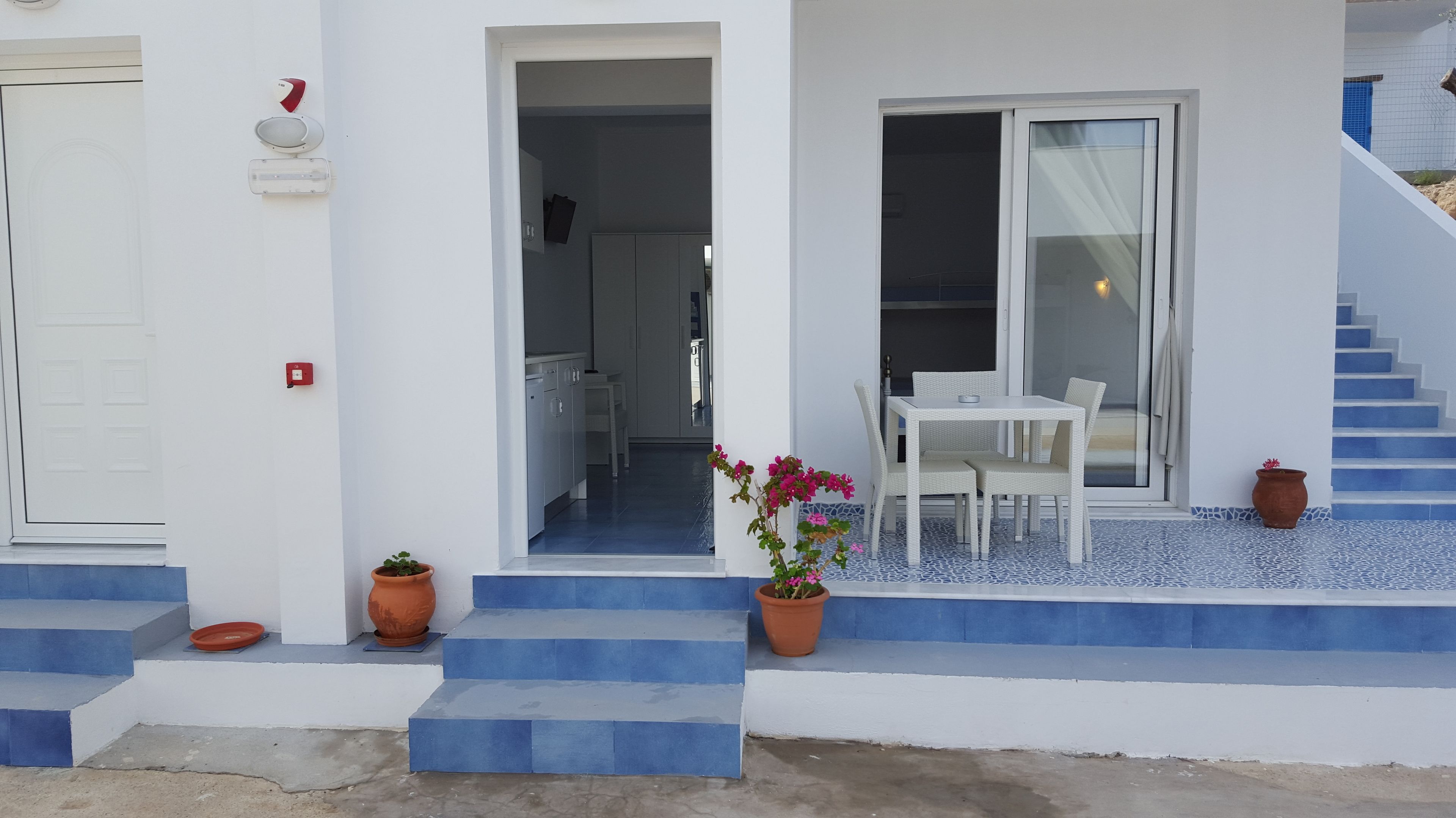Ein modernes Gebäude mit heller Fassade, blaue Stufen zu einer überdachten Terrasse und einer Essgegend mit weißem Mobiliar.
