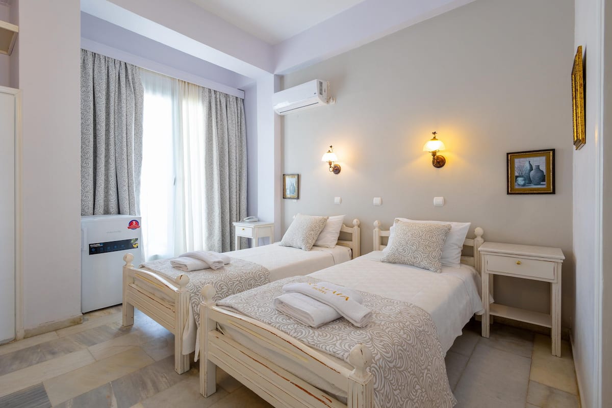 Una habitación de hotel con dos camas, una mesita de noche, aire acondicionado instalado en la pared, una ventana con cortinas y una lámpara fijada a la pared.