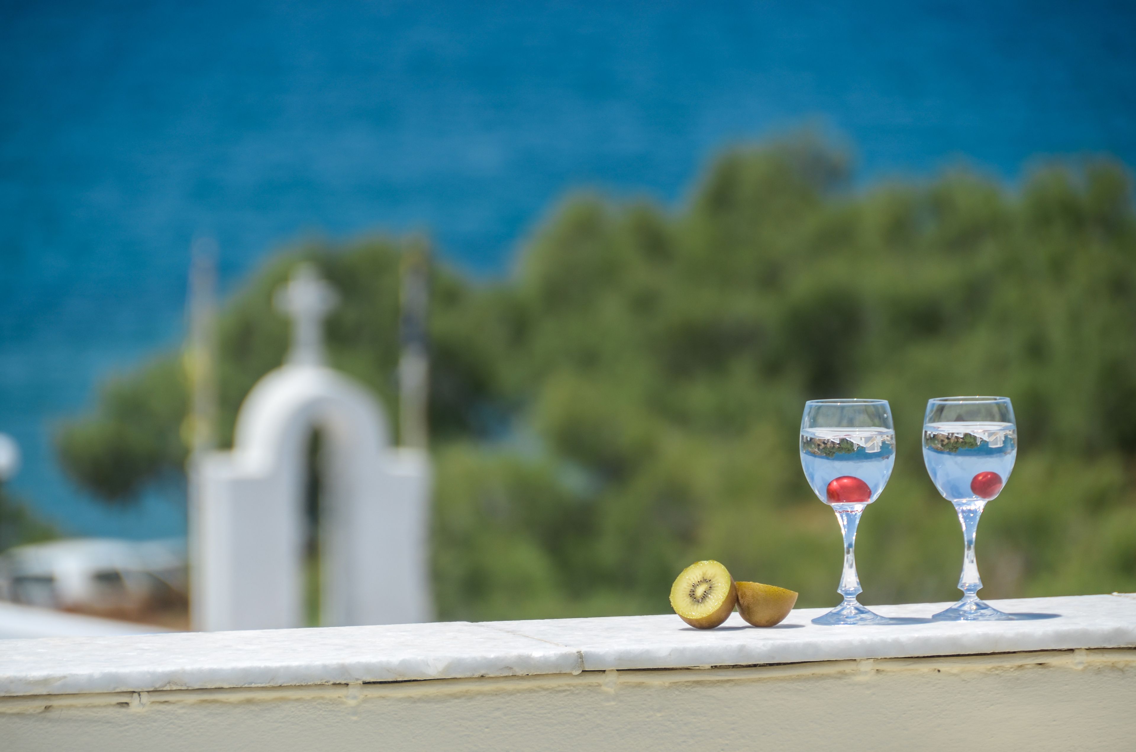 Two glasses of water with a kiwi and a cherry on a white ledge with a blurred background of greenery and a white structure.