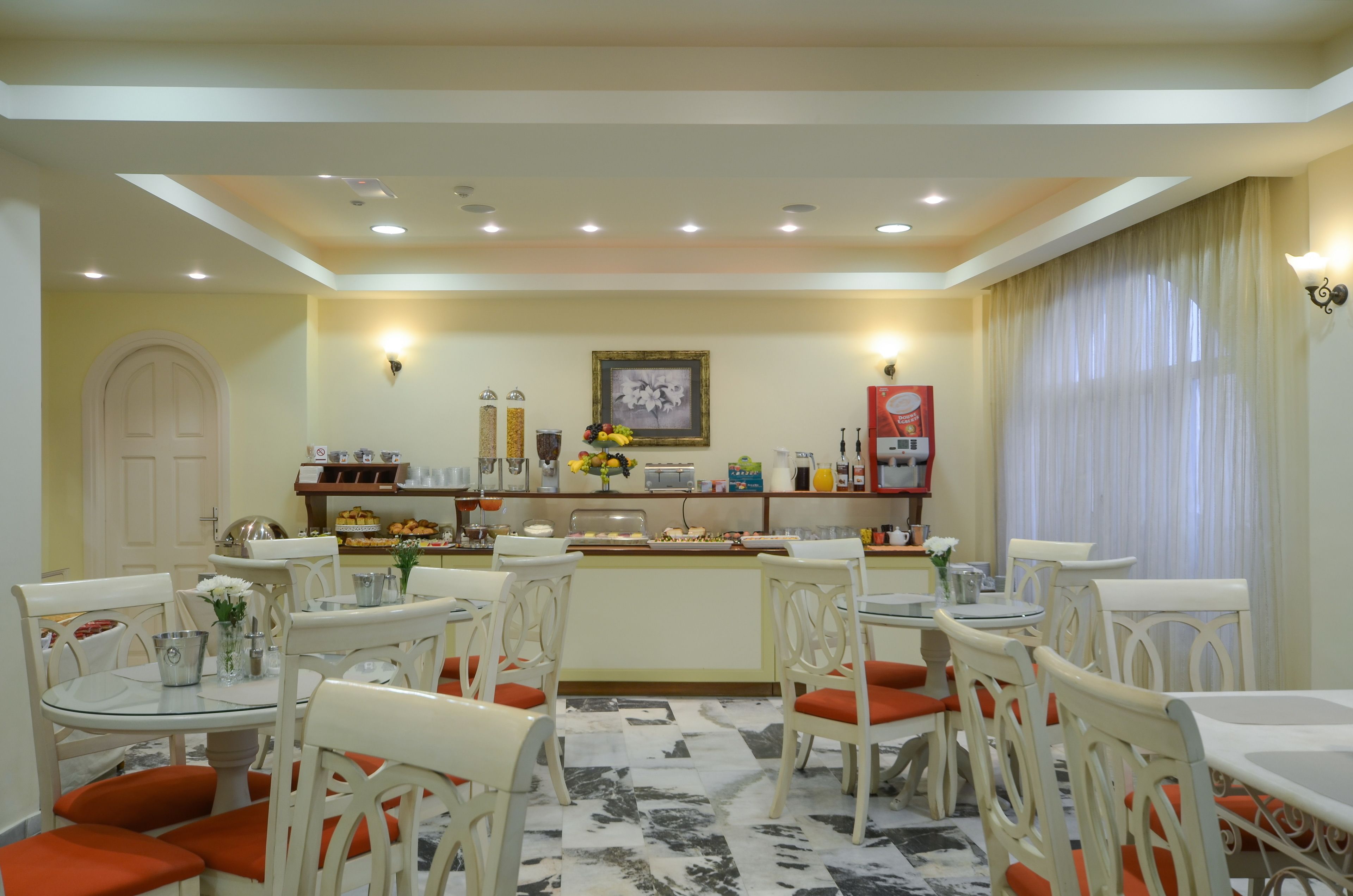 A dining area with white chairs and tables, a buffet counter with food and beverages, and a framed picture on the wall.