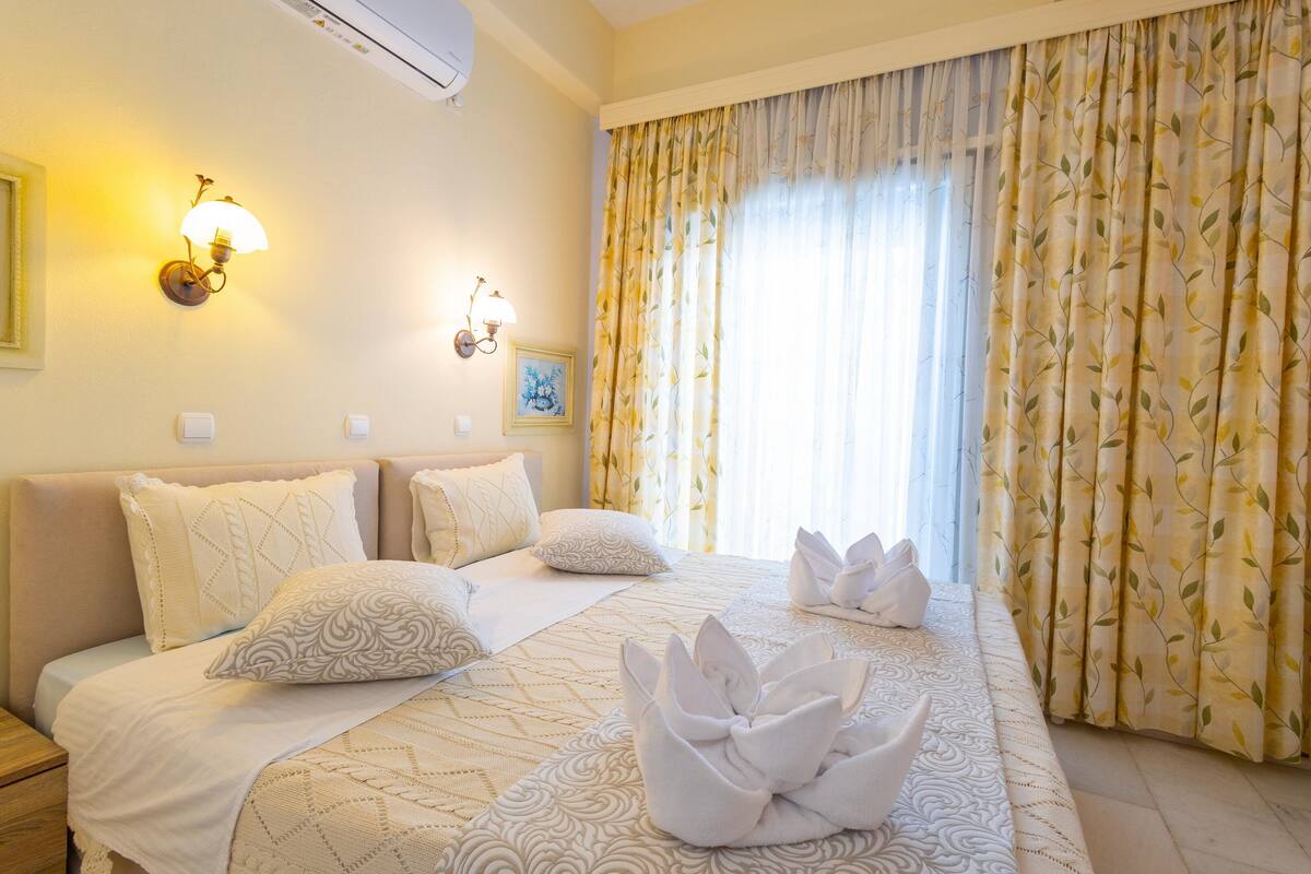 Habitación de hotel con cama, almohadas, lámpara de pared, cuadro enmarcado y ventana con cortinas.