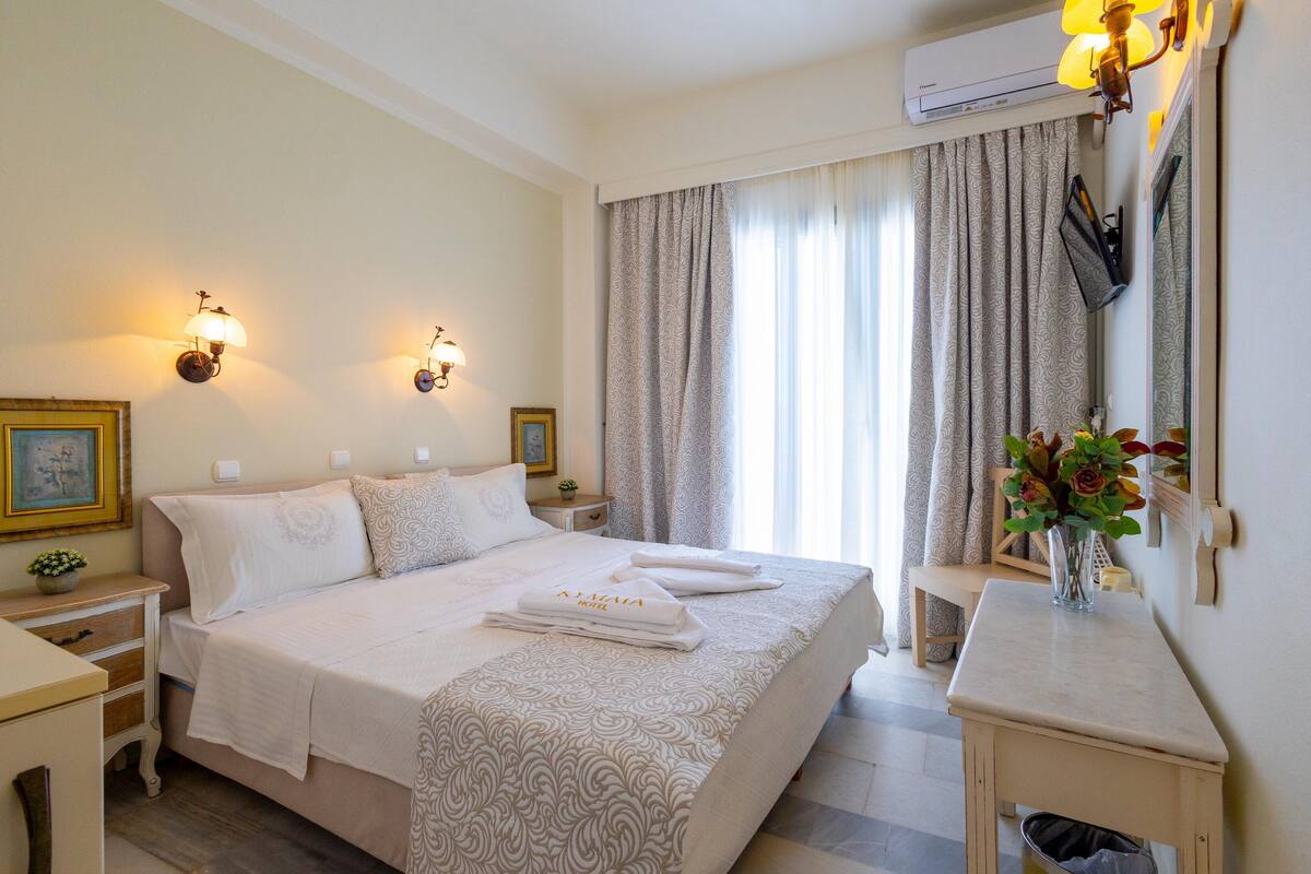Habitación de hotel con una cama bien hecha, dos mesitas de noche, una pequeña planta en maceta y un florero con flores.