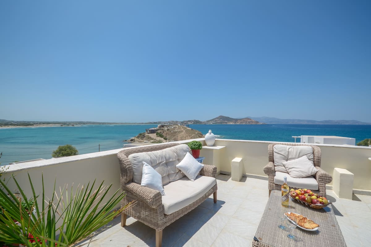 Una terraza con mobiliario de mimbre, una mesa con comida y bebidas, y vistas al mar y a las montañas.