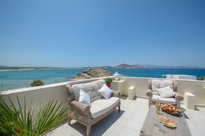 Una terraza con mobiliario de mimbre, una mesa con comida y bebidas, y vistas al mar y a las montañas.