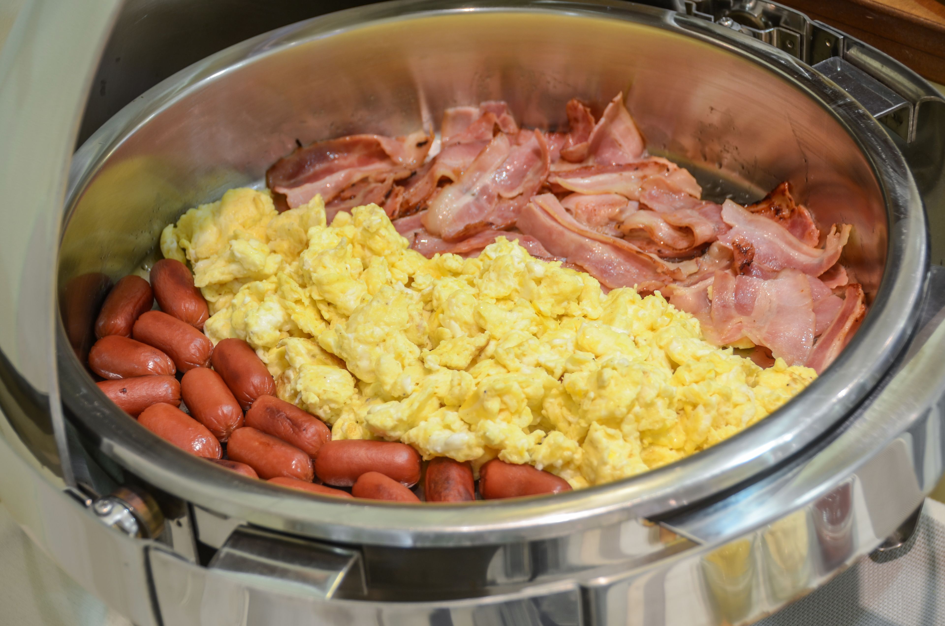 A buffet with scrambled eggs, bacon, and hot dogs.