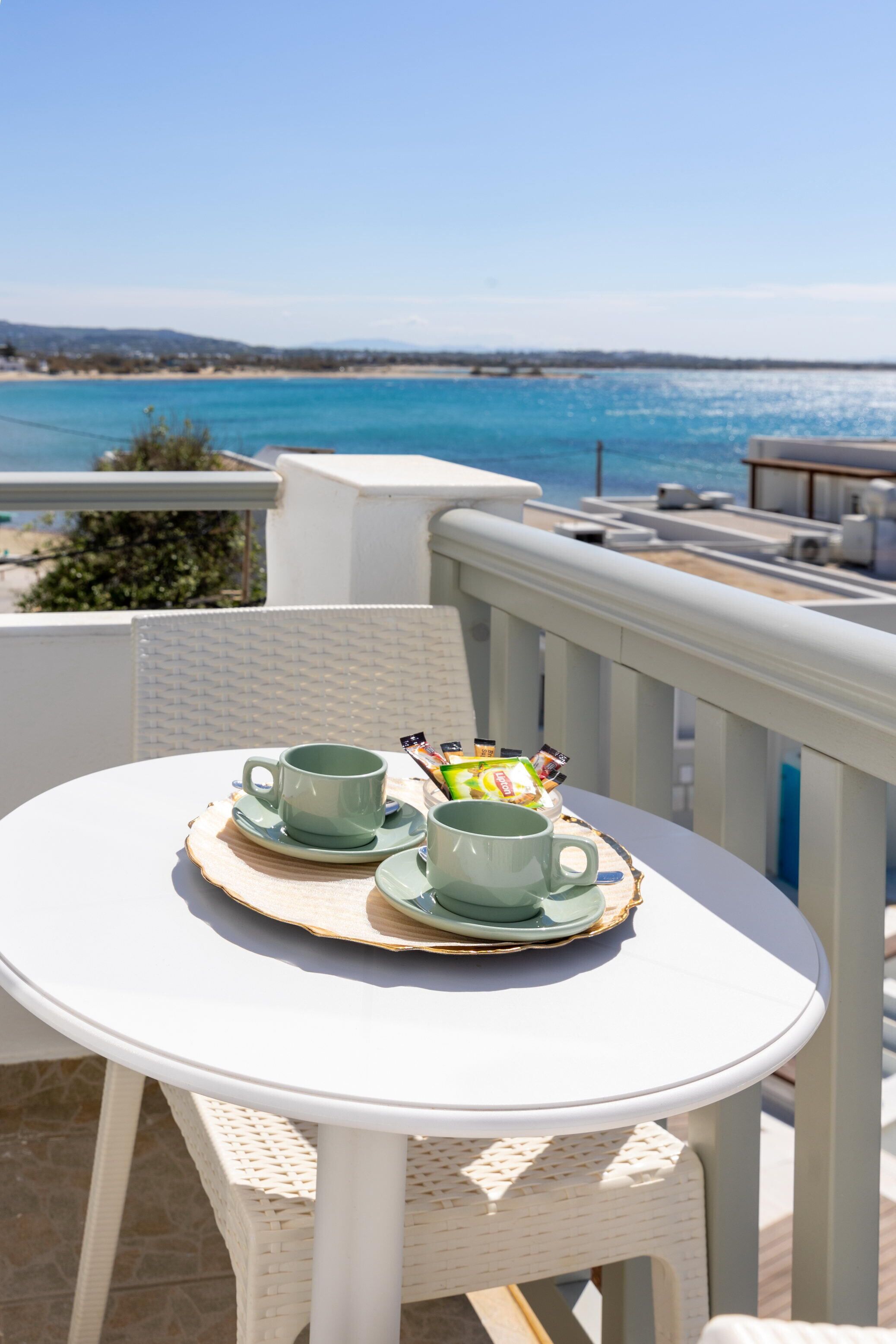 A round white table with two green cups and a tray of snacks on a balcony overlooking a beach.