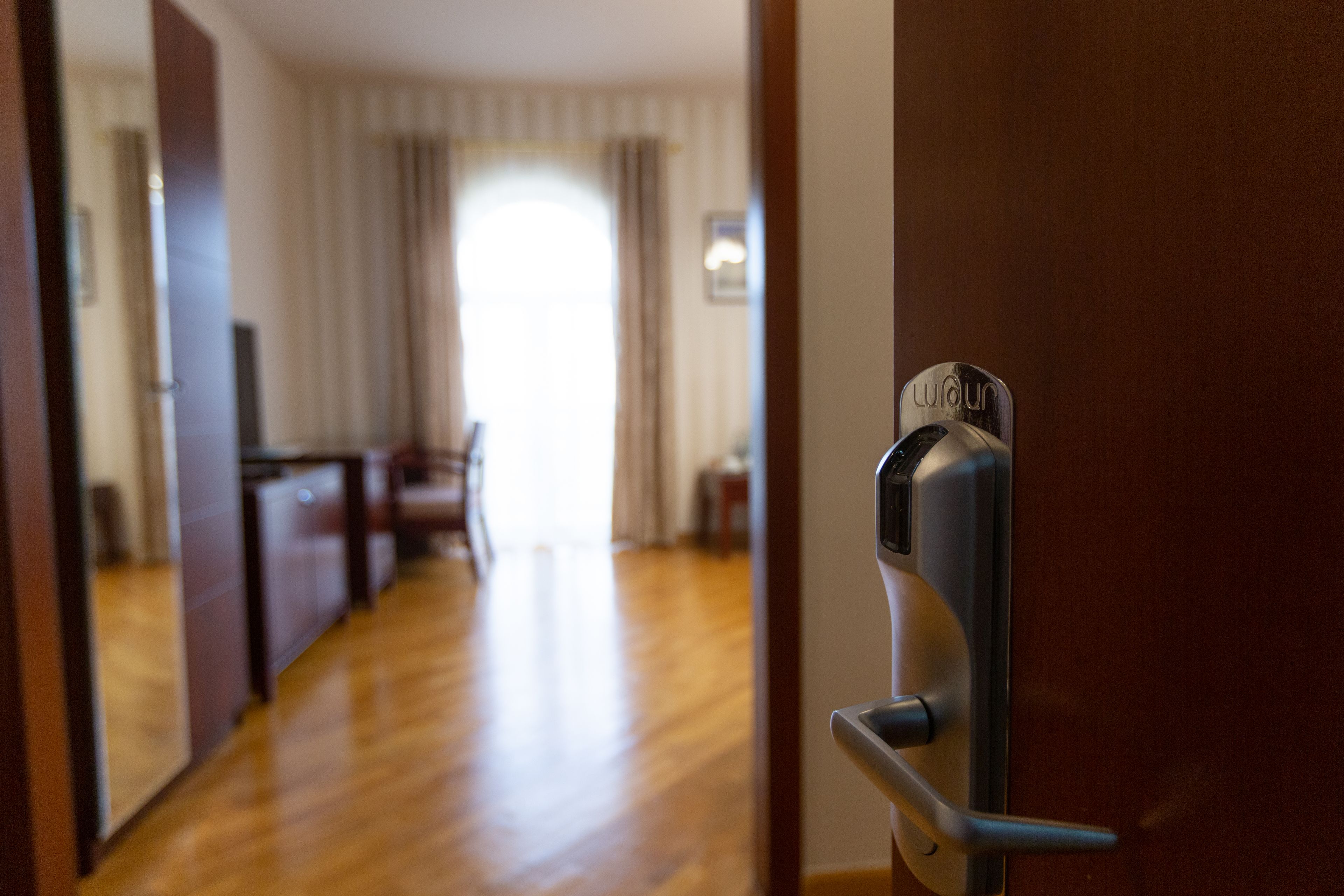 Entrada de una habitación de hotel con suelo de madera, una puerta con lector de tarjeta de acceso y vista a un comedor al fondo.
