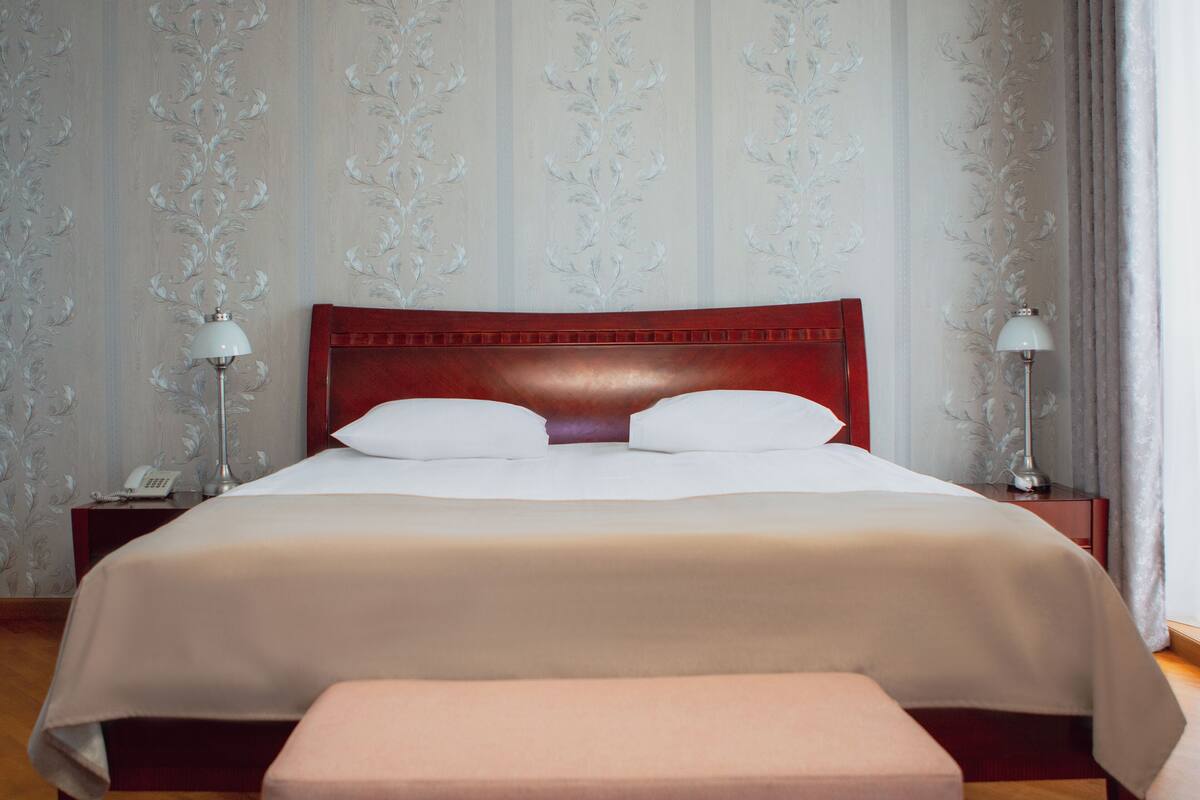 Cama bem arrumada com cabeceira de madeira, duas luminárias de cabeceira e uma mesinha com um telefone.